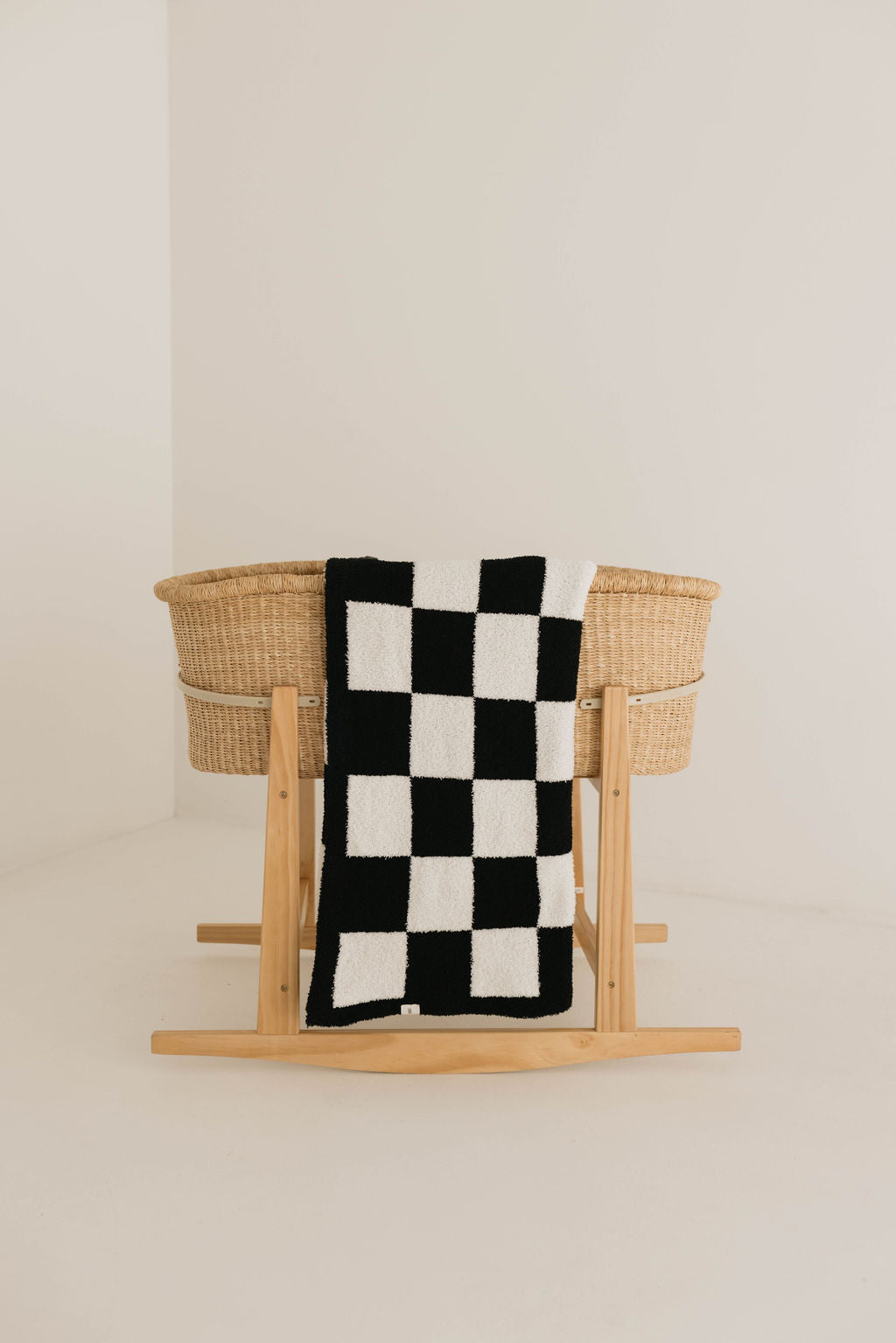 A woven bassinet stands in a minimalist room. Draped over one side is a Plush Blanket in a black and white checkerboard pattern by Forever French Baby, certified to the OEKO-TEX Standard 100. The overall setting features neutral colors and simple decor, highlighting the natural texture of the woven bassinet and the bold pattern of the soft blanket.