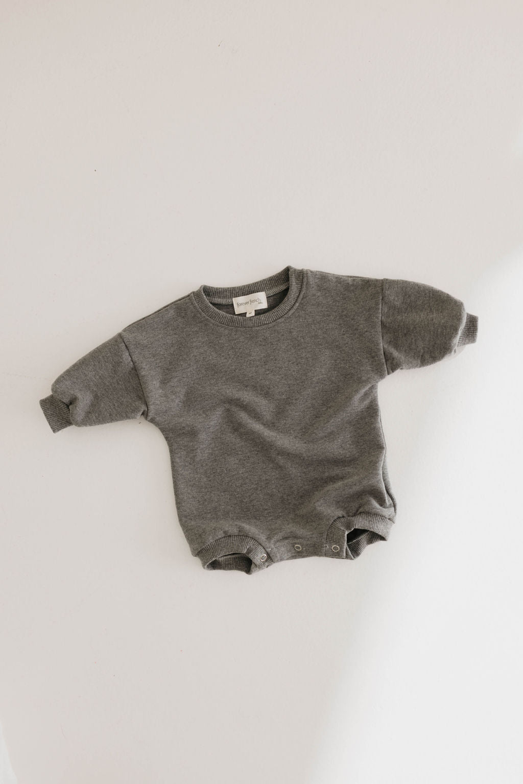 The Forever French Baby Sweatshirt Romper in charcoal grey is displayed flat on a light-colored surface. This long-sleeve romper features a round neck, ribbed cuffs, and convenient snap buttons around the leg openings for easy changing.