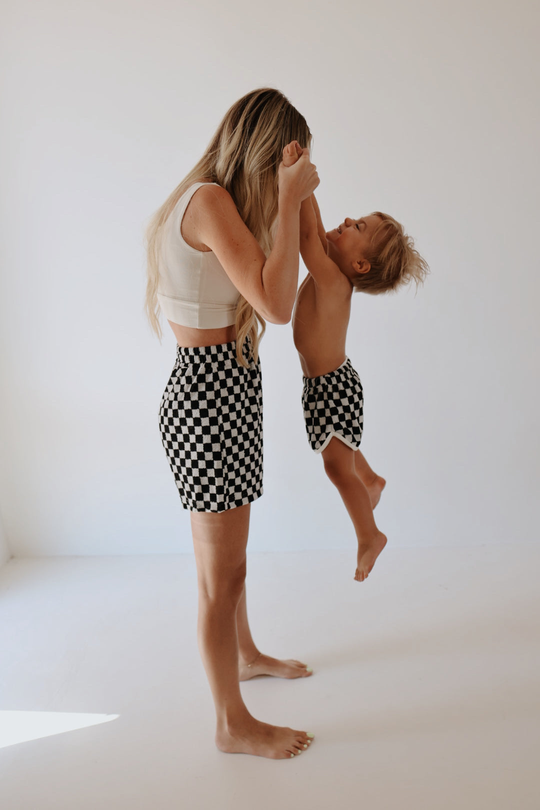 A woman with long hair wearing a white top and Women's Terry Shorts in Black + White Checkerboard by forever french baby is holding the hands of a young child dressed in matching checkered shorts, lifting them playfully off the ground in a light-filled, minimalist room. Both are barefoot and smiling joyfully.