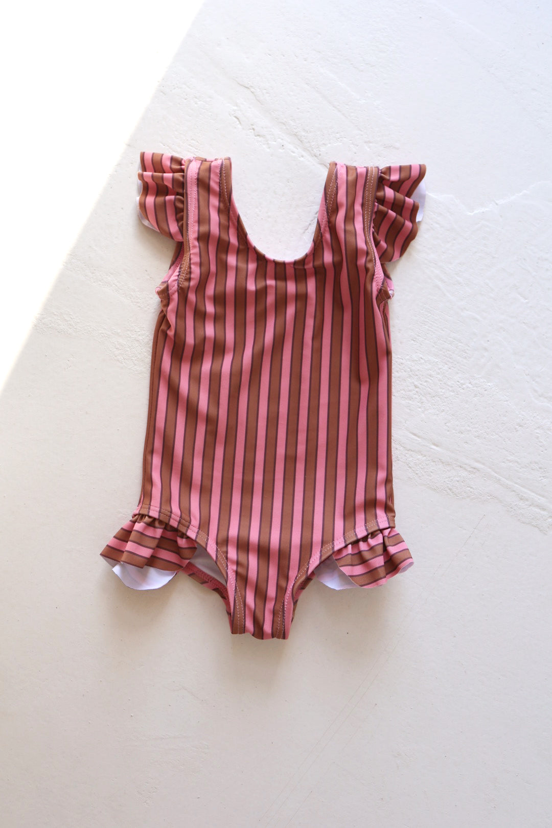 The One Piece Girls Swimsuit | Boardwalk from forever french baby, showcasing pink and brown earth-tone stripes, features flutter sleeves, a scoop neckline, and ruffled leg openings, is displayed on a white surface.