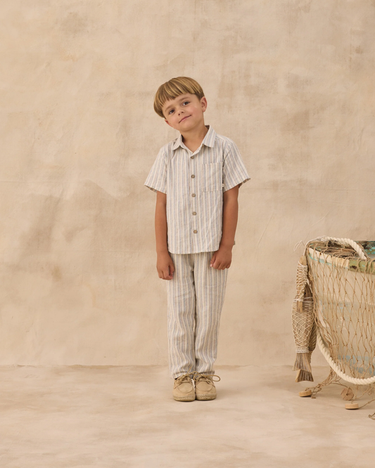 A young boy with light brown hair stands on a beige floor wearing the Rylee + Cru Ethan Trouser in Nautical Stripe, paired with a matching top. He leans to one side, with a basket and textured wall in the background.