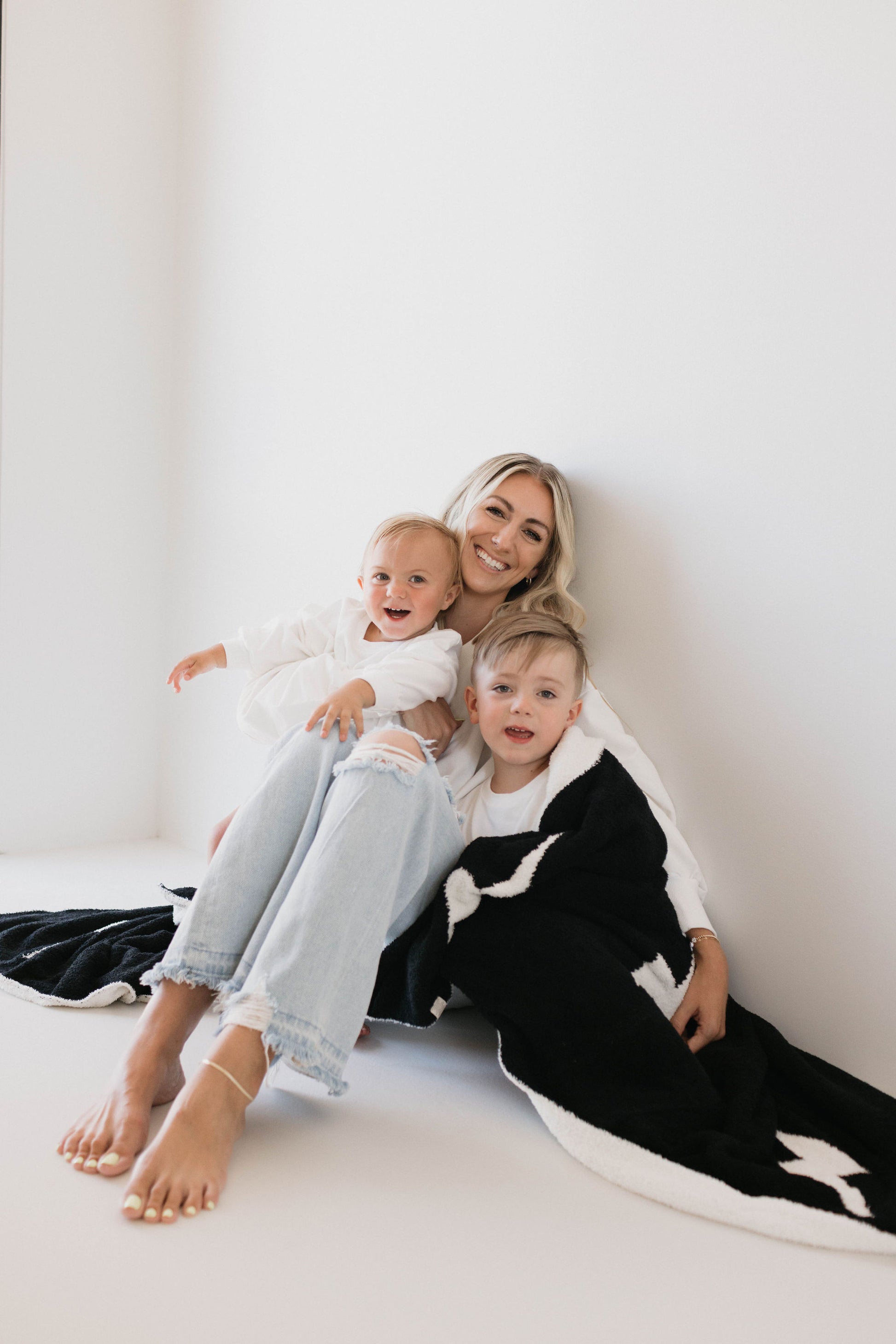 A woman smiles while sitting on the floor next to a white wall with two young children. One child sits on her lap with an open-mouthed smile, and the other, wrapped in a dreamy soft "Plush Blanket | Black & White Lightning Bolt" from forever french baby made of 100% Microfiber, is nestled beside her. All three are dressed in light-colored clothes.