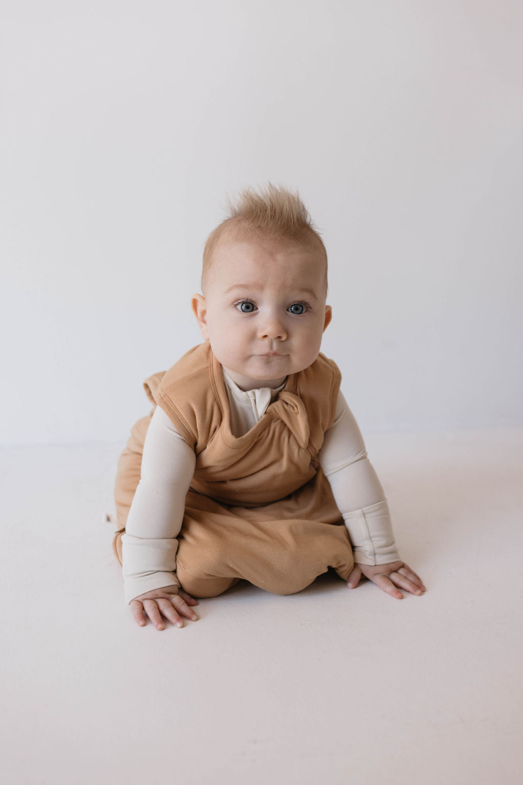 A baby with light skin and short blonde hair sits on the floor, wearing a tan outfit over a long-sleeved cream shirt and the eco-friendly Bamboo Sleep Sack | Desert Sunset by forever french baby. The baby has a neutral expression with a plain white wall in the background.