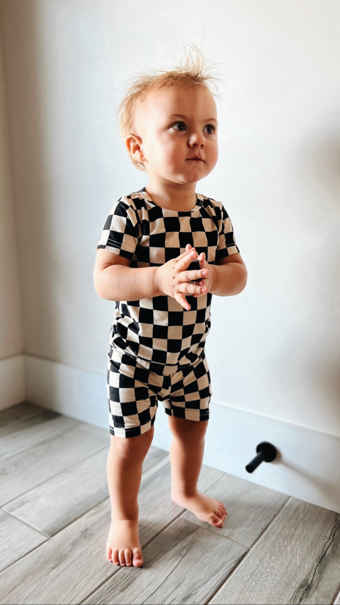 A toddler stands barefoot on a wooden floor, dressed in the Bamboo Short Set | Black Checkerboard by forever french baby. The child has light hair and gazes slightly upward and to the side with both hands held together in front. The background is a plain off-white wall.