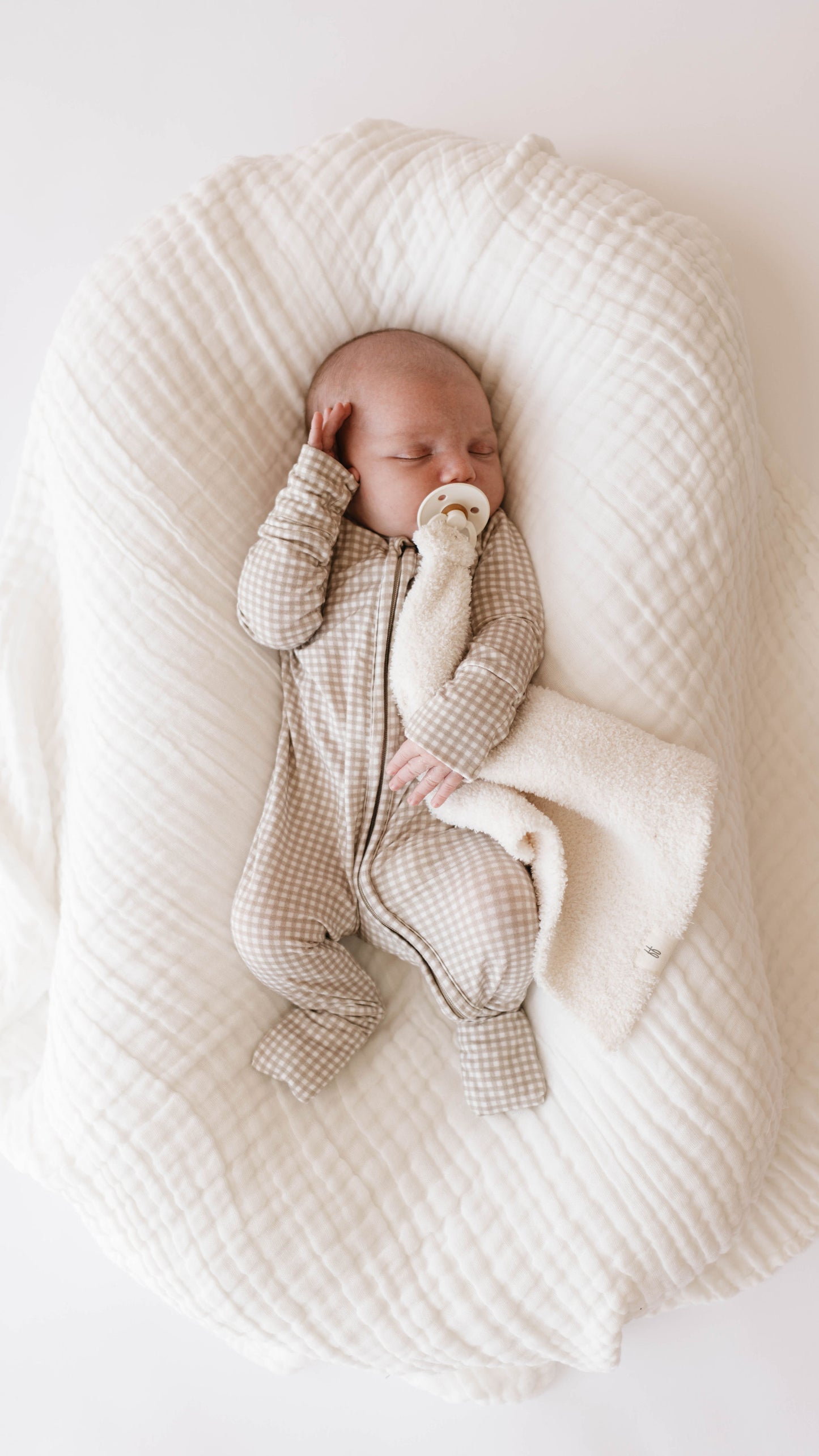 A baby sleeps on a cozy, quilted white blanket, adorned with the Crib Sheet | Cloud from Forever French Baby. The baby is wearing a zip-up, checkered onesie from the same brand. A pacifier rests in its mouth as it holds a small, soft blanket close. One hand is near its head while the other grips the blanket. The scene is serene and peaceful.