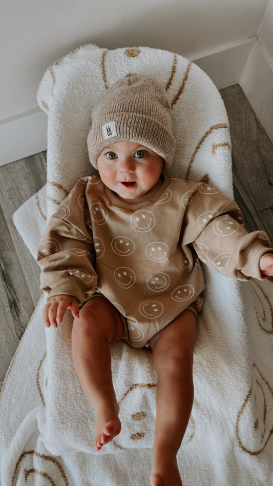 A baby with big blue eyes and a happy expression wears the forever french baby Just Smile Long Sleeve Romper—a tan organic cotton piece with white smiley faces—paired with a beige knit beanie, perfect for showcasing adorable style.