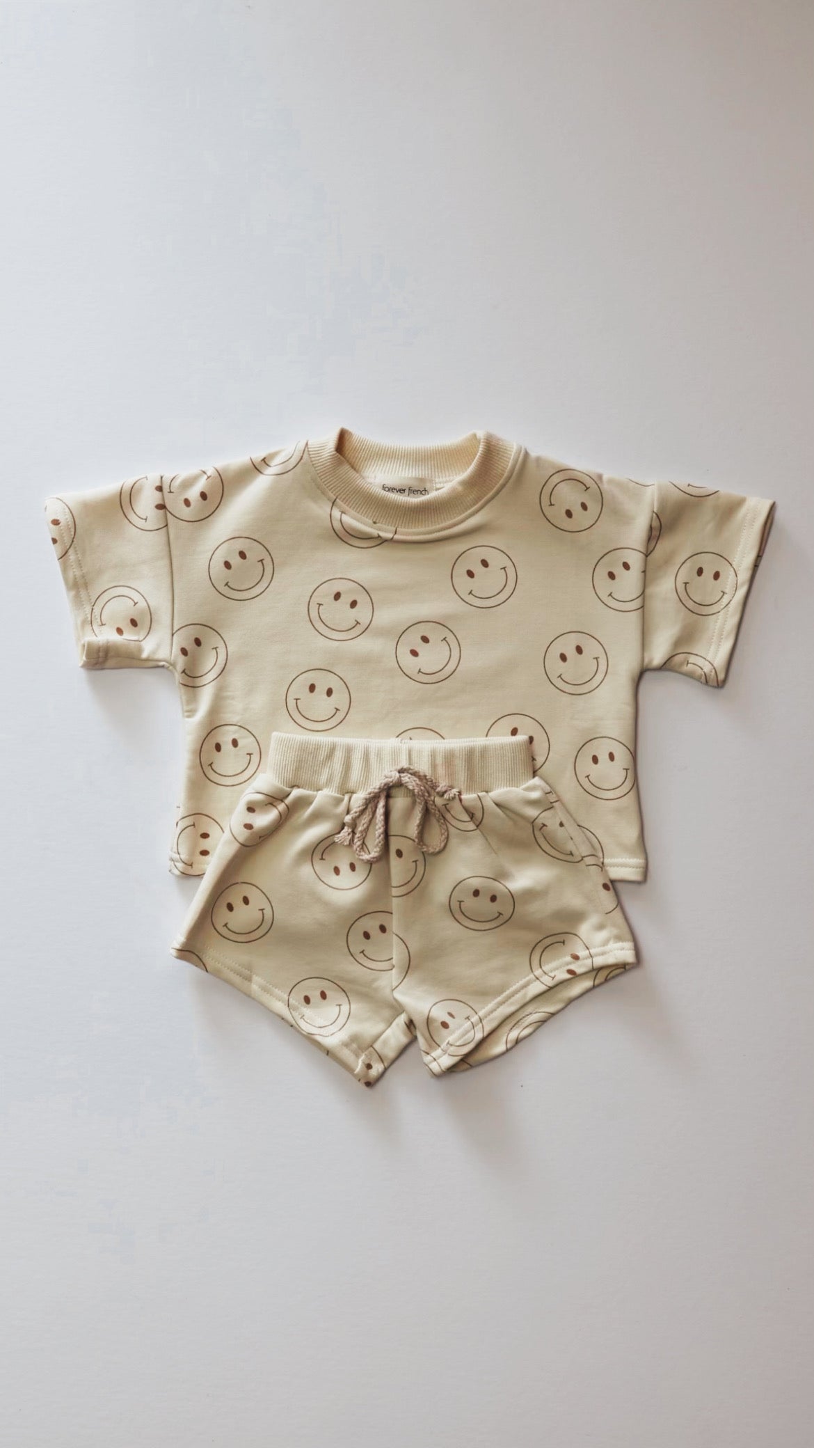 The Short Set | Just Smile Ivory from forever french baby is a delightful children's outfit that includes a short-sleeved shirt and matching shorts, both decorated with a charming pattern of smiling faces. This ivory set is showcased laid out flat against a plain white background, making it an ideal piece for versatile wardrobe transitions.