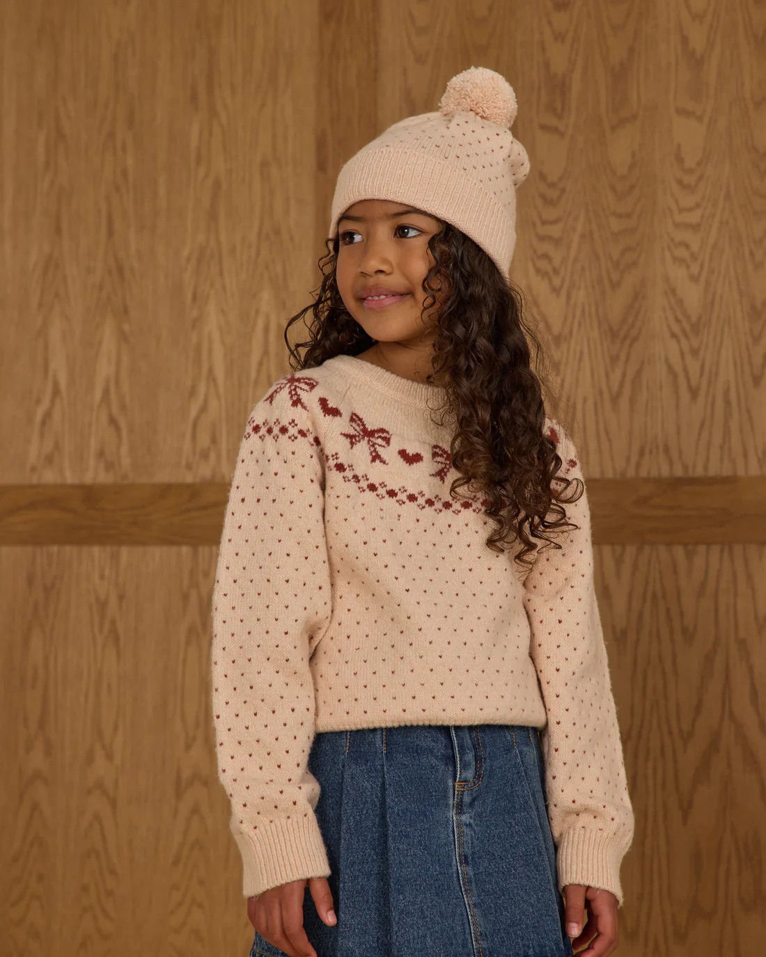 A young girl with long curly hair stands in front of a wooden wall, smiling as she looks to the side. She wears the Rylee + Cru Fair Isle Sweater | Bows—a cozy knit sweater featuring red designs—paired with a denim skirt and a warm knit hat.
