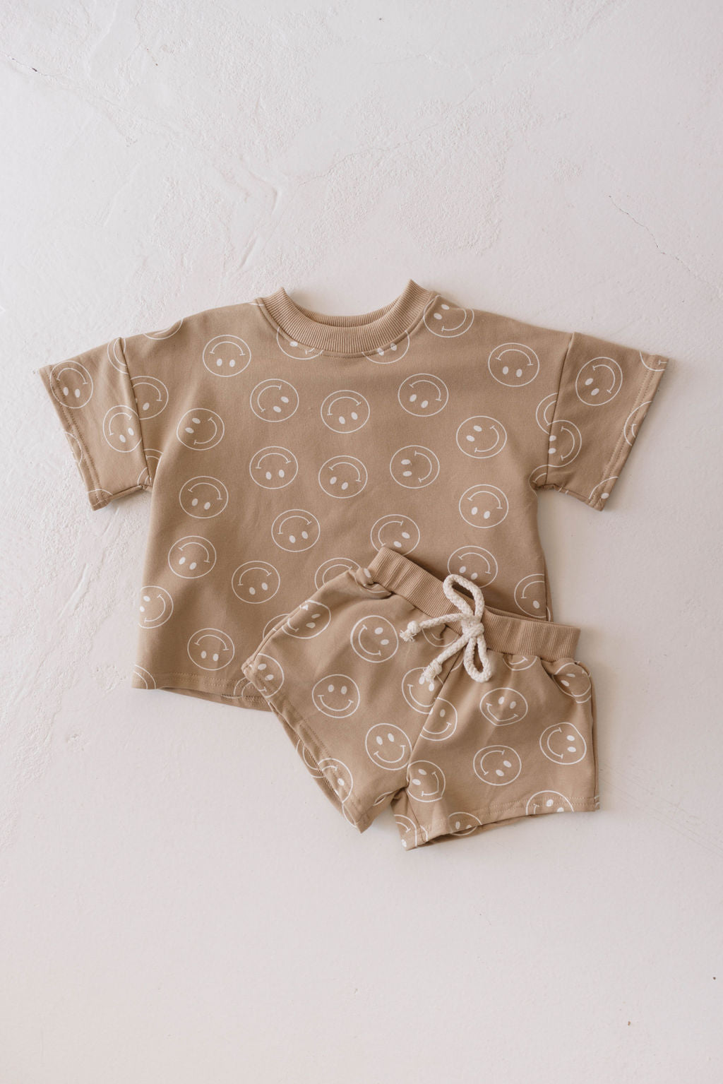 A delightful addition to any children's wardrobe, this Just Smile Tan short set by forever french baby features a short-sleeved shirt and shorts with drawstrings, both adorned with white smiley face patterns. The ensemble is charmingly displayed on a white textured surface.