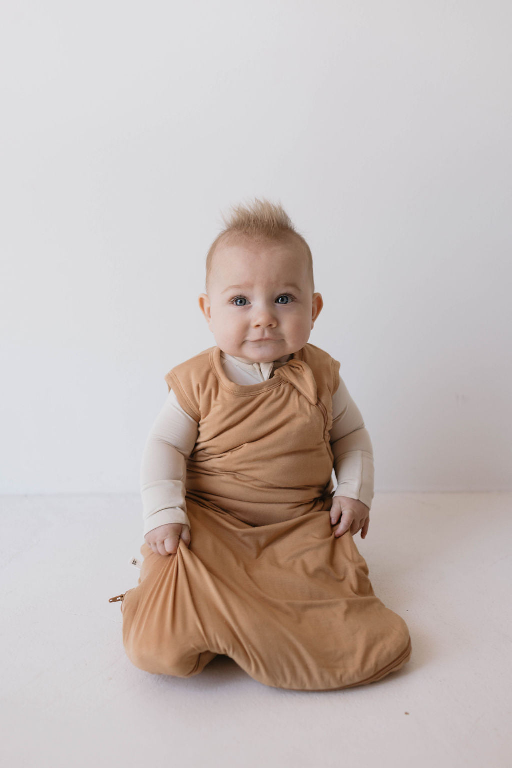 A baby with light brown hair sits on a white background, wearing the Bamboo Sleep Sack | Desert Sunset by forever french baby over a long-sleeved white shirt. The baby looks at the camera with a slight smile, holding onto its eco-friendly wearable blanket with both hands.