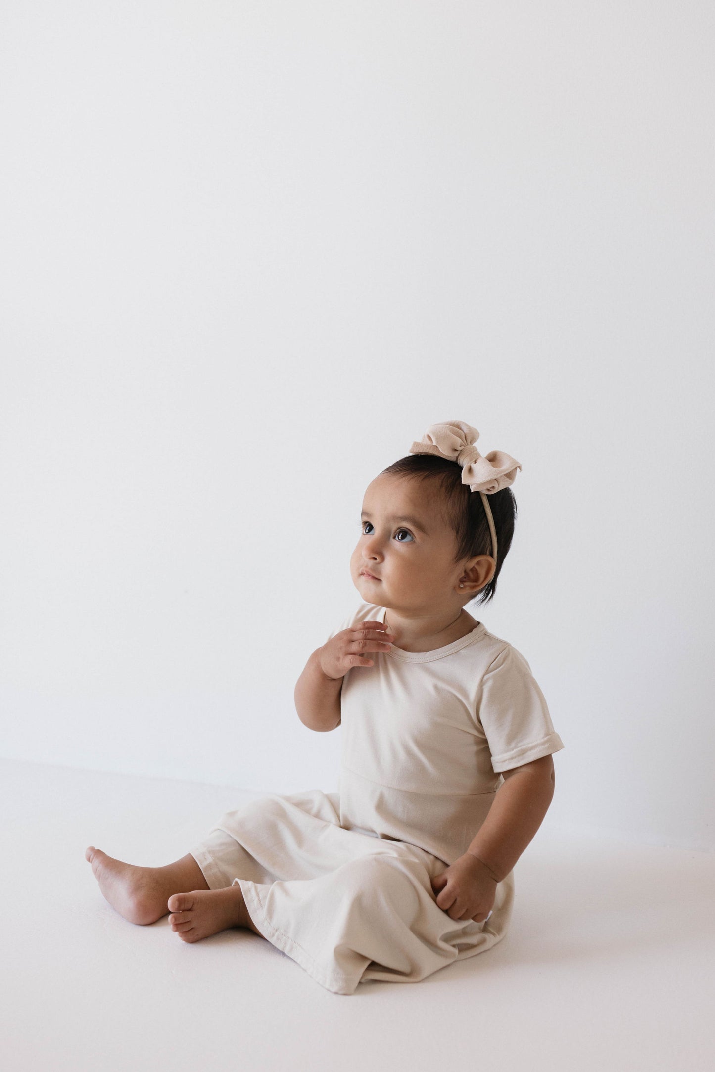 A baby wearing the forever french baby's Bamboo Dress in Oatmeal sits on the floor against a plain white backdrop, looking curious and charming with a soft pink bow adorning their head.