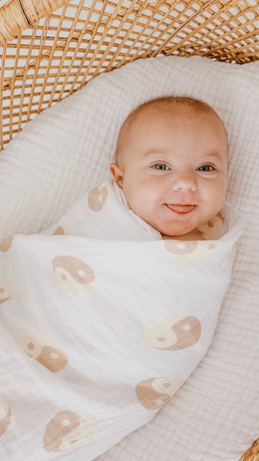 A baby with big eyes and a smile is swaddled in a "Muslin Swaddle | Tan & White Yin Yang" by forever french baby. The baby sits in a wicker bassinet on a soft, white, quilted surface.