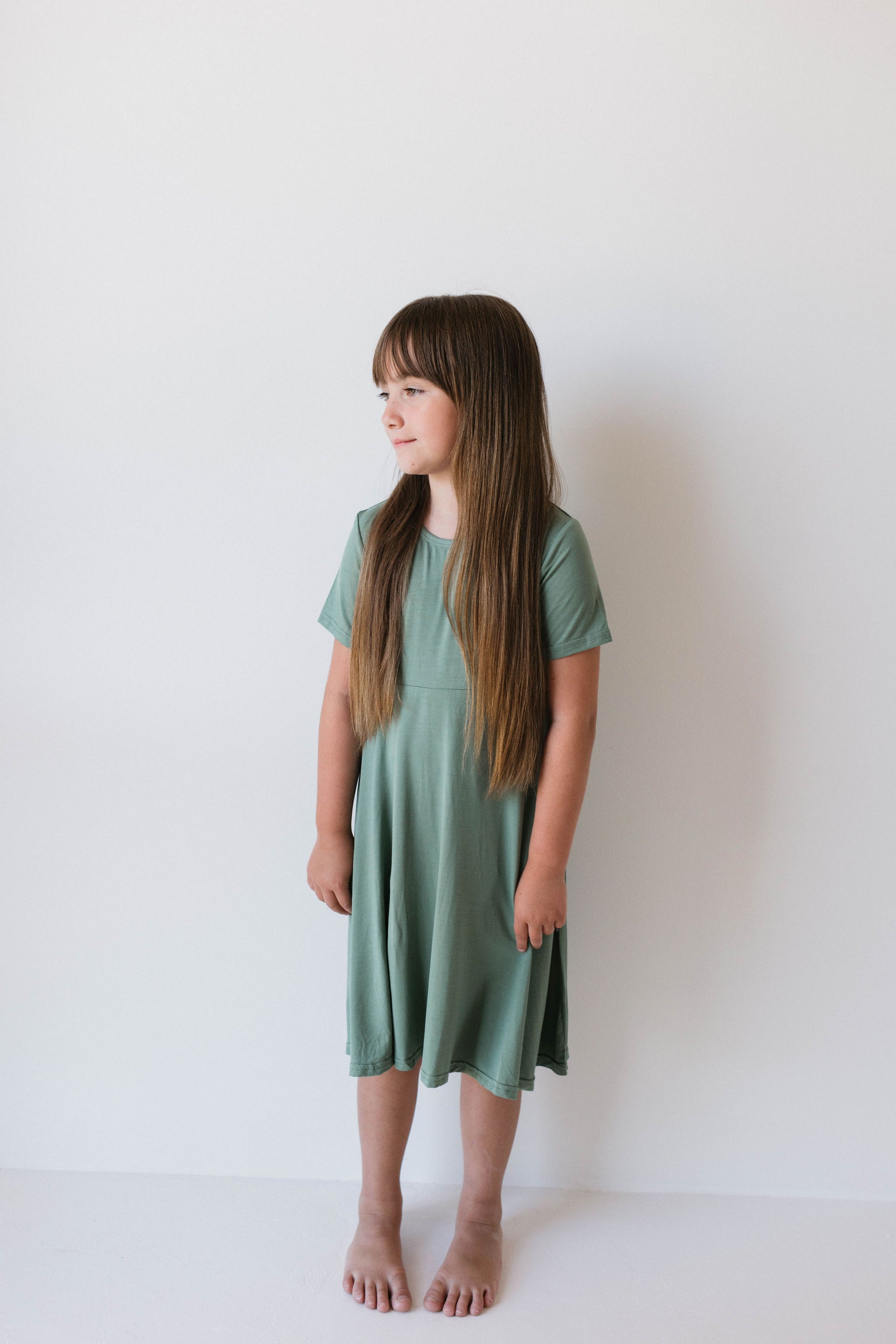 A young girl stands barefoot against a plain white background, looking to her left. She has long brown hair and wears the Bamboo Dress in Moss from Forever French Baby. She holds her hands together in front of her. This hypo-allergenic clothing ensures both comfort and style for sensitive skin.