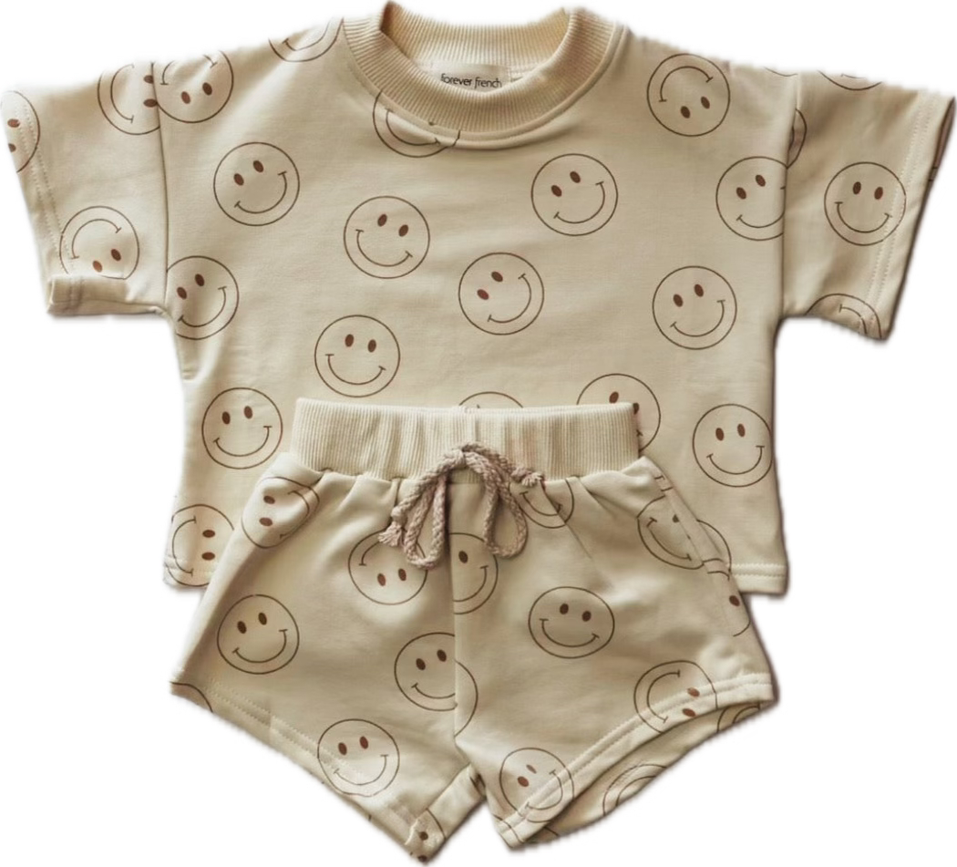 This delightful children's outfit from forever french baby, named the "Short Set | Just Smile Ivory," features an ivory short-sleeve top and matching shorts adorned with a pattern of simple, smiling face icons. The shorts have a drawstring waistband, making it an ideal transitioning piece for your little one's wardrobe.