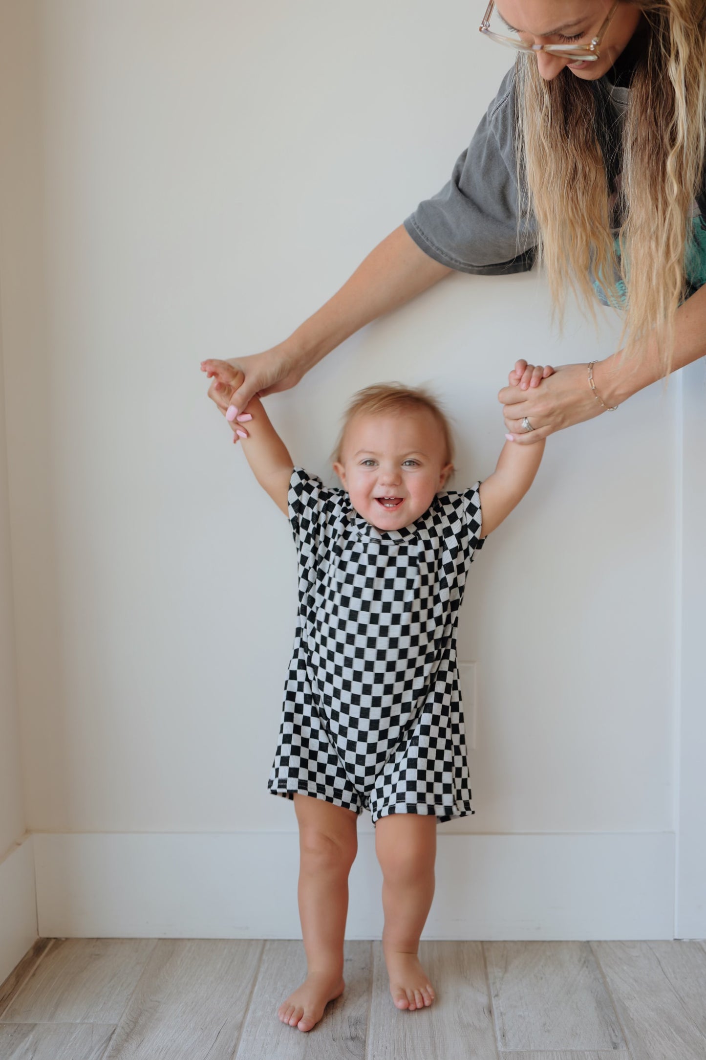 A young child in a Short Sleeve Ribbed Romper | Black & Grey Checkerboard from forever french baby is smiling while being held upright by an adult whose hands are gently supporting the child's raised arms. The background is a plain, light-colored wall and wooden floor.