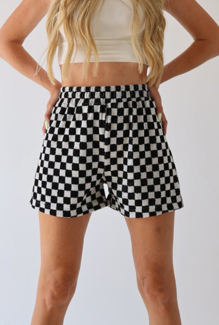 A woman with long hair is wearing a white cropped top and Women's Terry Shorts in Black + White Checkerboard by forever french baby. Her hands are placed on her hips, revealing her midriff against the plain white background, making for a perfect addition to any summer wardrobe.