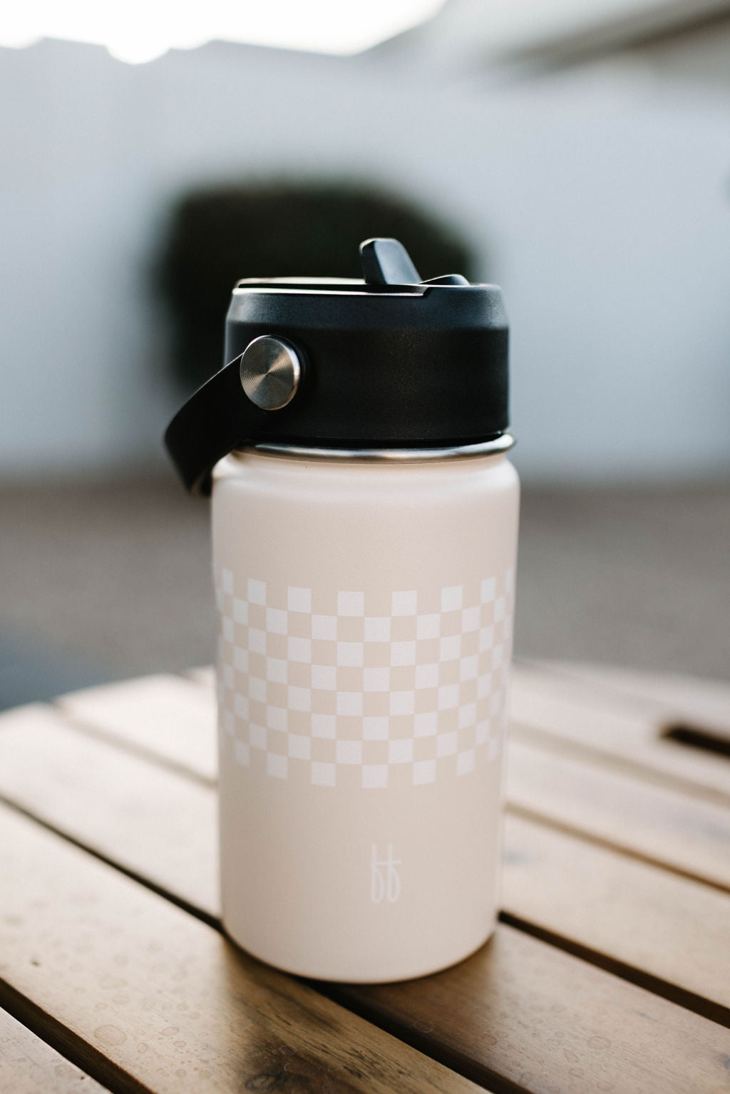 A tan ff Water Bottle from forever french baby, featuring a black cap and handle, sits on a wooden surface. The insulated BPA-free stainless steel bottle has a checkerboard pattern near the top and a stylized "bb" logo near the bottom. The background is blurred, showcasing a dark green bush and a white wall.