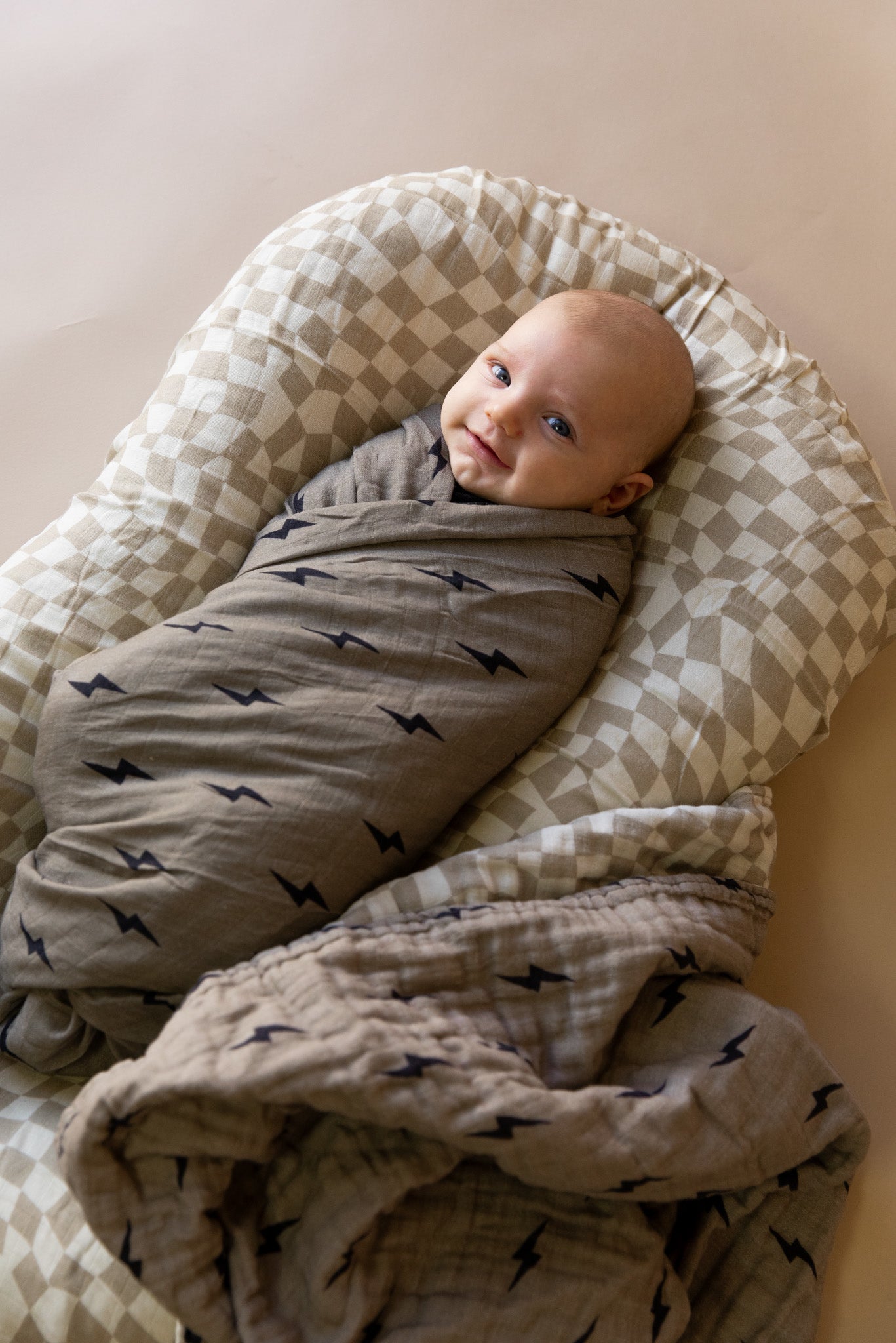 An infant is swaddled in a light gray Muslin Swaddle | Lightning Bolt by forever french baby, featuring small black lightning bolt patterns. The baby lies calmly on a cushioned, checkered pad with a soft beige background.