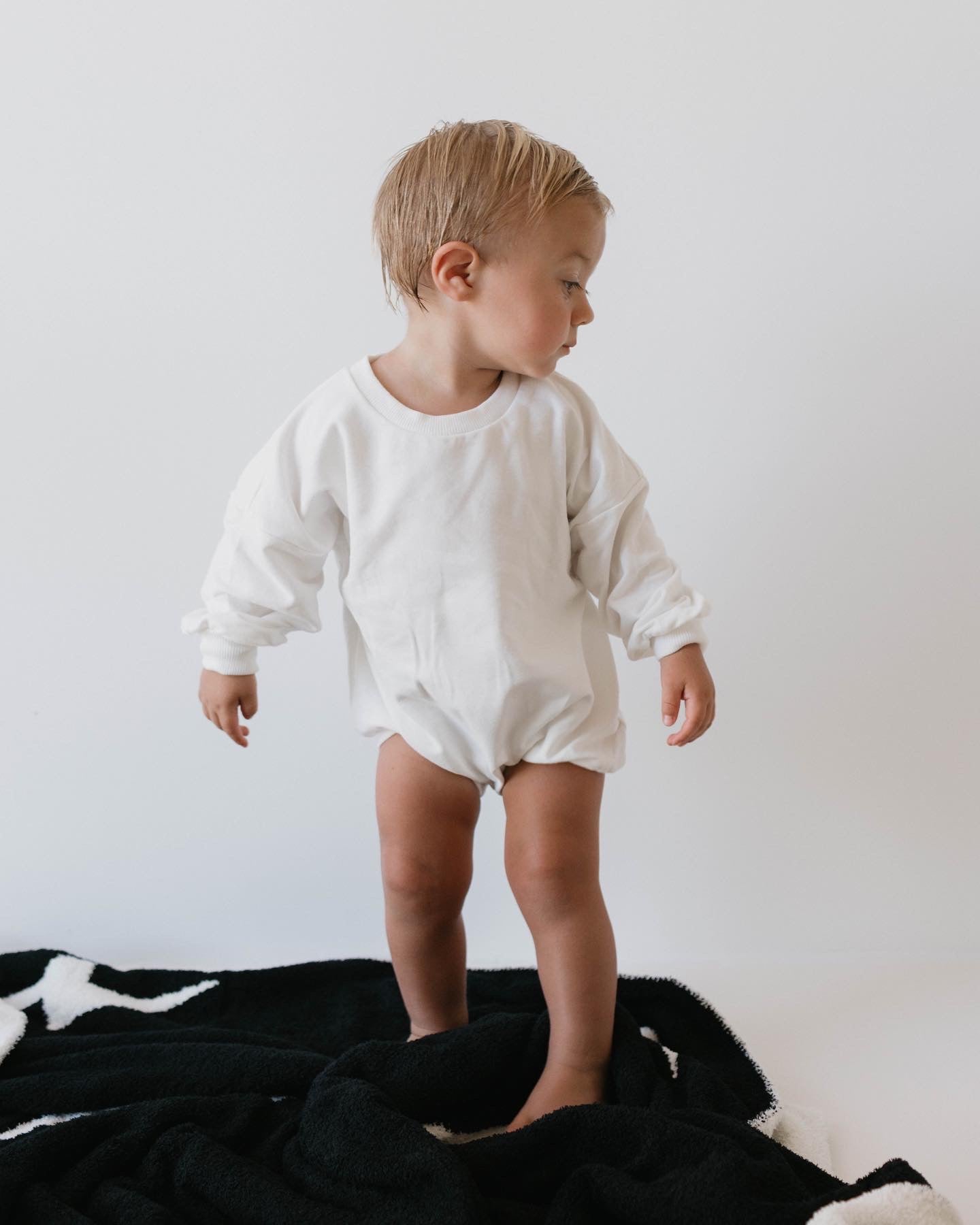 A young child with short blonde hair stands on a black and white blanket, dressed in the Dove Sweatshirt Romper by forever french baby. The child's onesie features convenient snap closures, and they are looking to the side against a plain white wall.