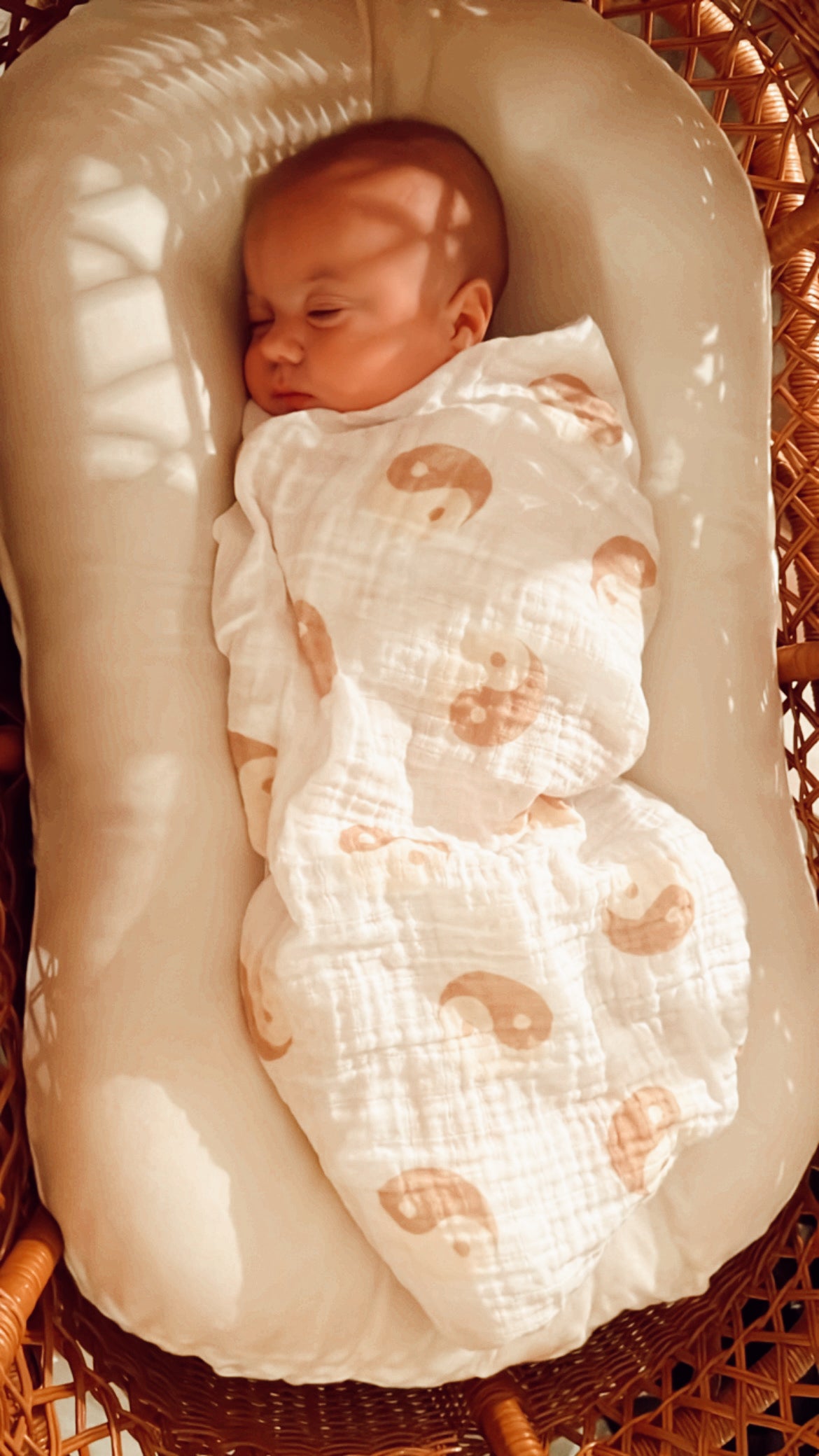 A forever french baby is peacefully sleeping in a cushioned wicker bassinet, swaddled in a Muslin Swaddle | Tan & White Yin Yang blanket from Forever French Baby. Soft sunlight filters through, casting gentle shadows.