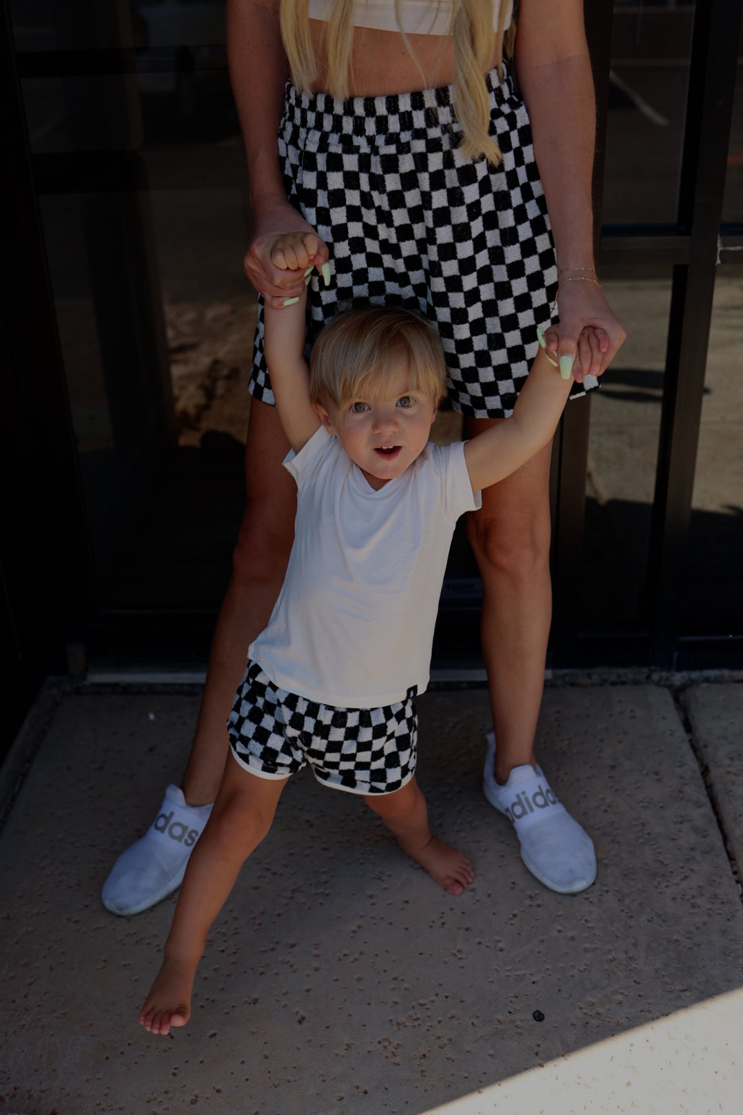 A woman holding hands with a small child standing in front of her, both showcasing their summer wardrobe in matching forever french baby Women's Terry Shorts in Black and White Checkerboard. The child sports a white T-shirt, while the woman pairs a crop top with white slip-on shoes.