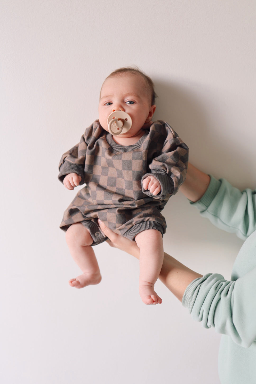 A baby wearing the "Sweatshirt Romper | Faded Brown Checkerboard" from forever french baby with a pacifier in their mouth is held up against a white wall by an adult hand in a green sleeve. The baby looks calm and is positioned facing the camera.