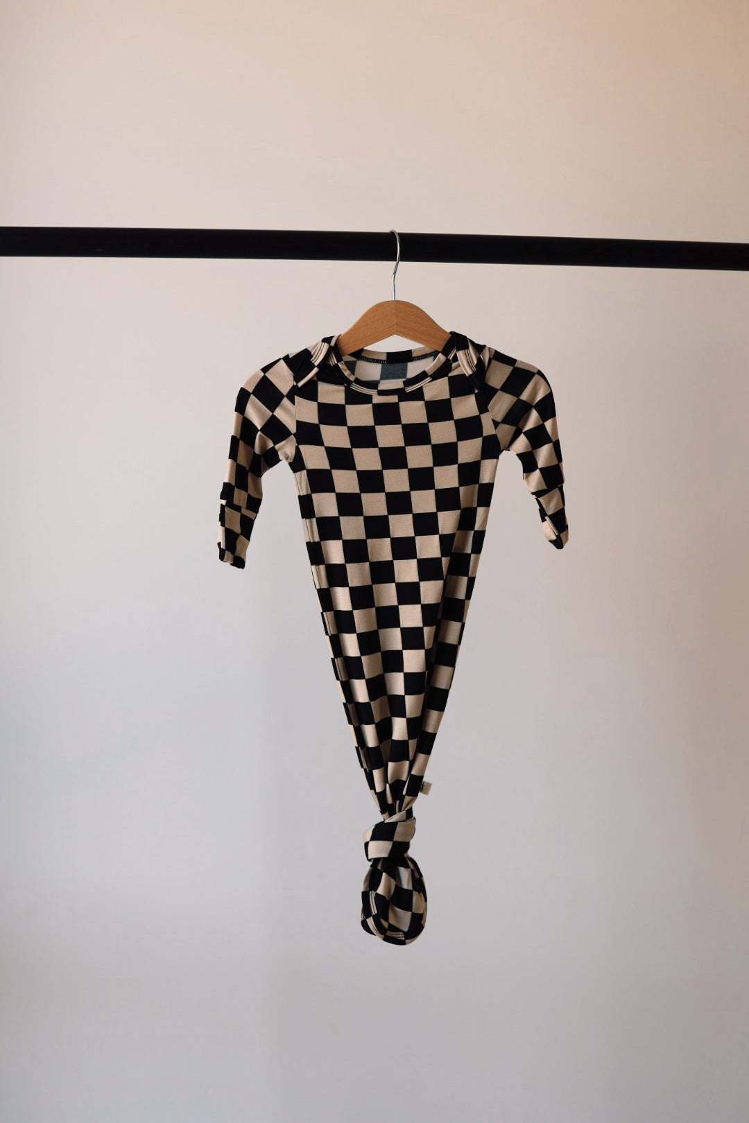 A newborn essential, the "Knotted Bamboo Gown | Black Checkered" from forever french baby, hangs on a wooden hanger. This hypo-allergenic, long-sleeve garment is knotted at the bottom and displayed against a plain, light-colored background on a thin, black clothing rod.