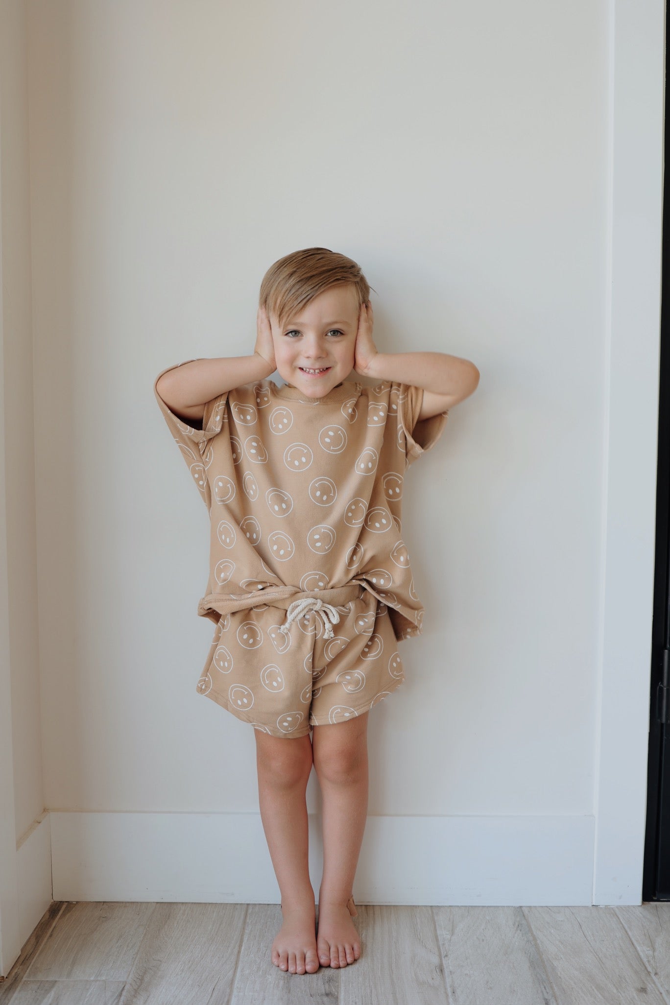 A child with short blonde hair stands barefoot against a white wall, smiling and covering their ears with their hands. They are wearing the "Just Smile Tan" short set from forever french baby, both pieces adorned with a white smiley face print.