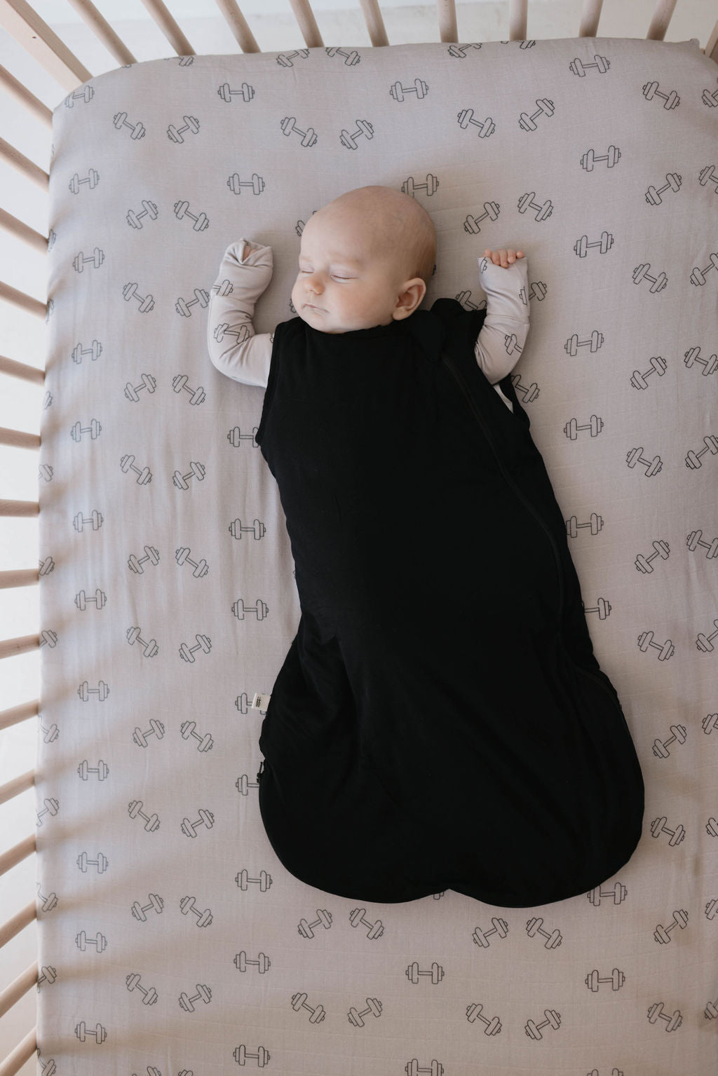 A baby sleeps peacefully on their back in a crib, wrapped in a Midnight Black Bamboo Sleep Sack by forever french baby and wearing a long-sleeved shirt with a cute pattern. The crib has a soft, light-colored sheet decorated with small barbell illustrations, while the wooden bars provide delicate security.