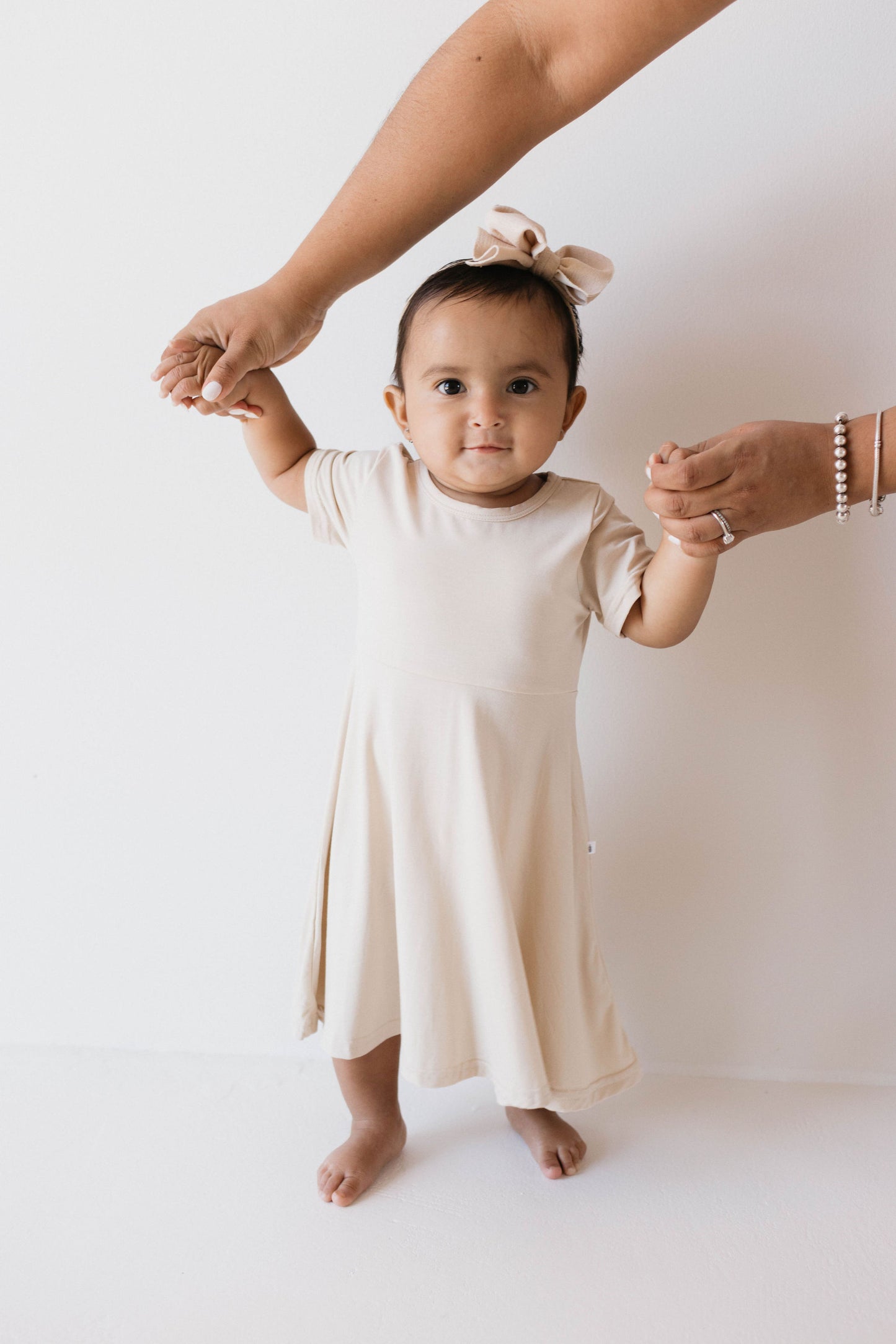 In a photo against a plain white background, a baby wears forever french baby's Oatmeal Bamboo Dress and matching bow headband. The baby stands with an inquisitive look, supported by an adult's hands from above, showcasing the charm of breathable bamboo clothing.