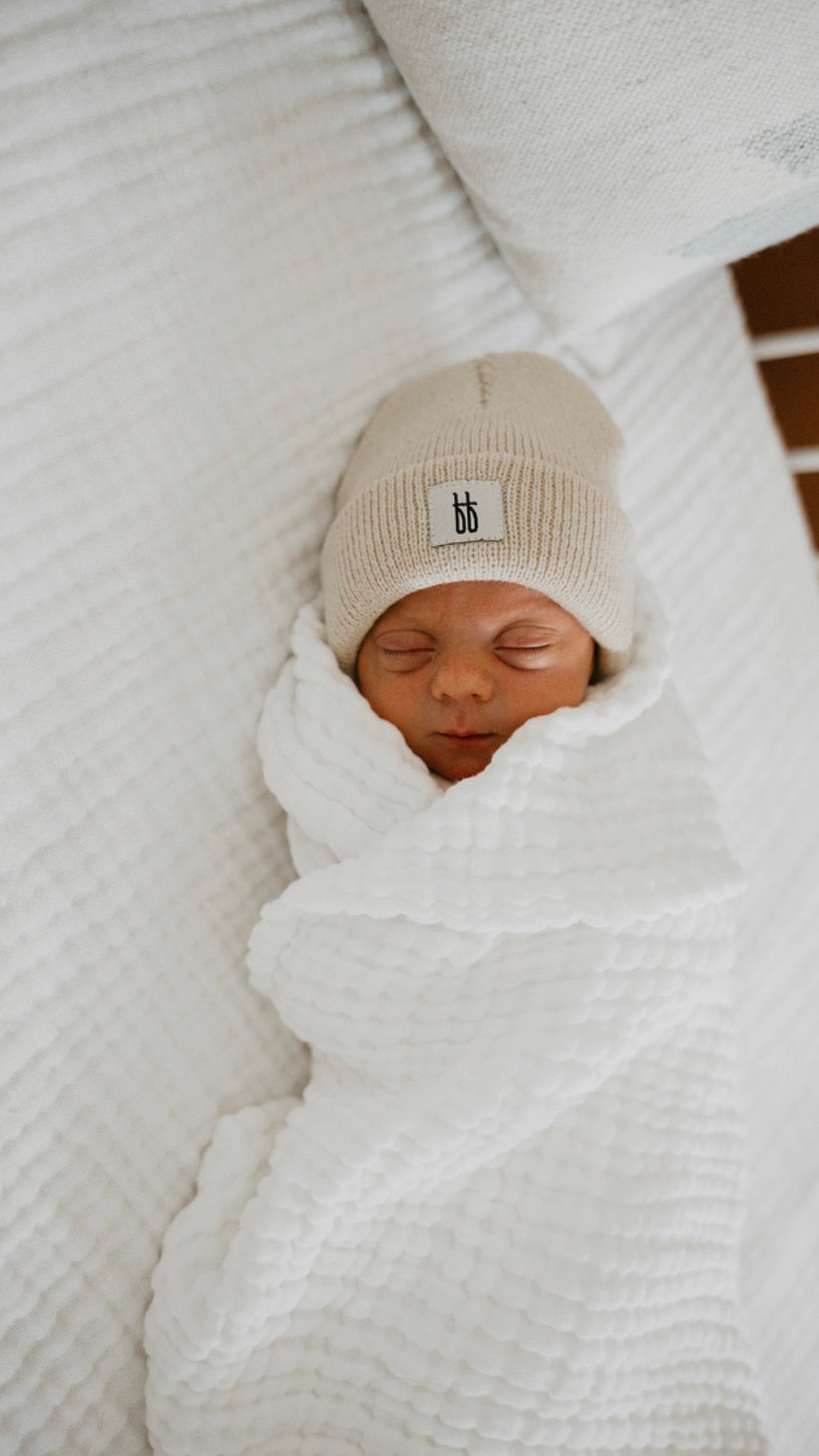 A newborn baby from the Forever French Baby collection is swaddled snugly in a white, textured blanket and lies on a pristine Crib Sheet | Cloud. The baby wears a beige knit beanie, sleeping peacefully with eyes closed. The setting appears calm and cozy. Remember to wash with like colors and tumble dry on low.