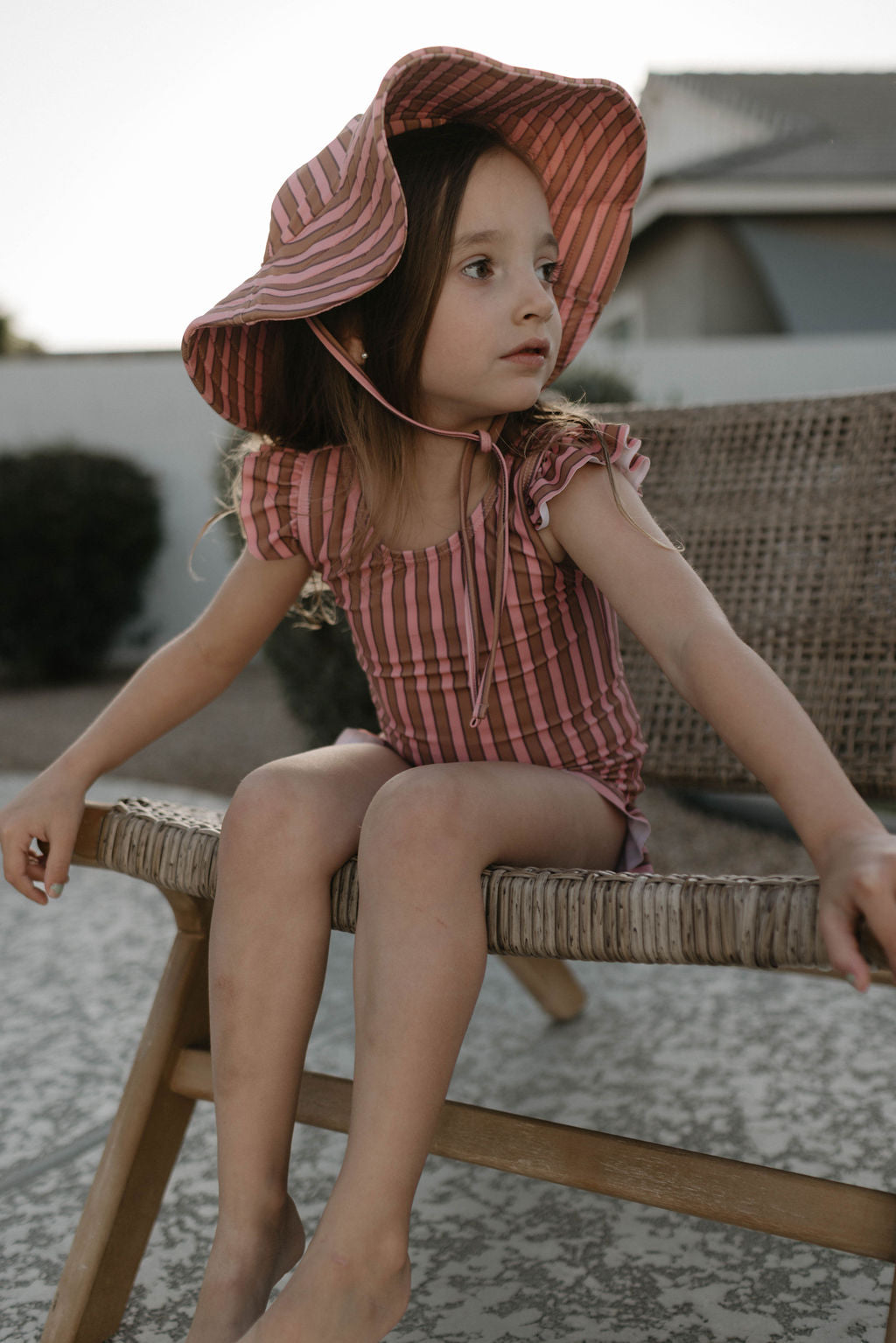 A young girl wearing a forever french baby One Piece Girls Swim Suit in the Boardwalk pattern sits on a wicker chair outdoors. She appears to be looking at something off to the side. The background includes a building and some greenery, suggesting the setting is likely a patio or outdoor area.