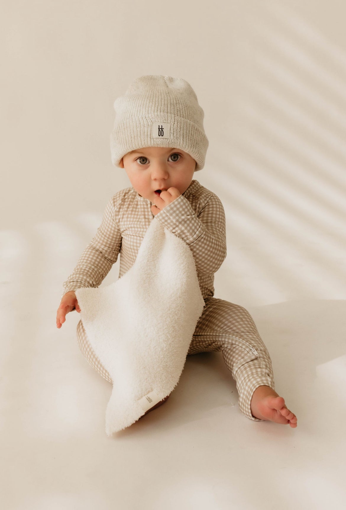 A baby is sitting on the floor wearing a beige checkered outfit and a white knit beanie, holding a forever french baby Lovey in Cream made of microfiber feather yarn. The baby has a focused expression, and the background is softly lit with shadows creating a gentle striped pattern.