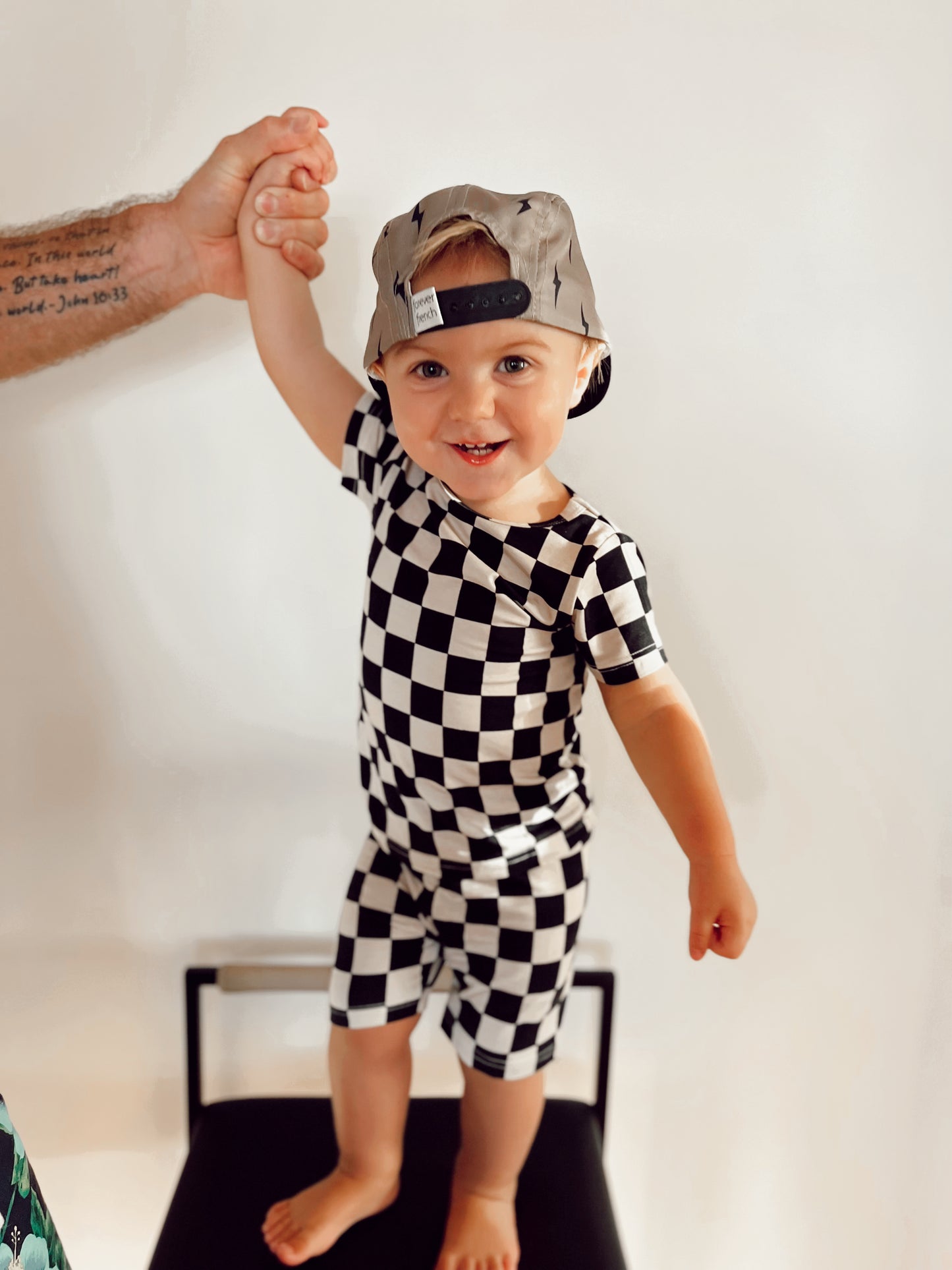 A cheerful toddler stands on a chair holding an adult's hand, dressed in the Bamboo Short Set in Black Checkerboard by forever french baby. The child wears a gray baseball cap backwards, set against a plain, light-colored background.