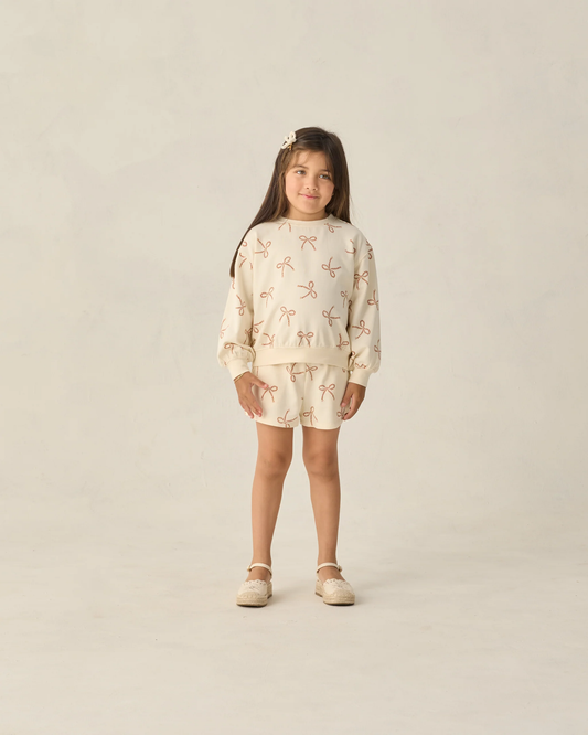 A young girl stands against a light beige background, wearing the Rylee + Cru Summer Terry Set | Sailor Bows—featuring a relaxed sweatshirt and matching shorts with a subtle bow pattern. She has long brown hair, a hair clip, and beige shoes.