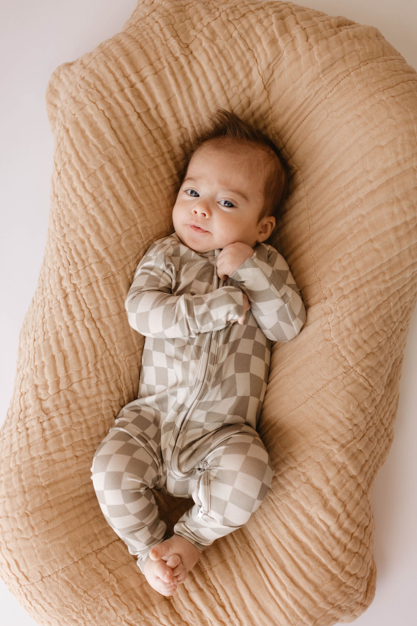 An infant, clad in a beige and white checkered onesie, lies on a soft, textured light brown cushion with the Latte Quilt by forever french baby. The baby wears a calm expression with their hands lightly clasped near their face.