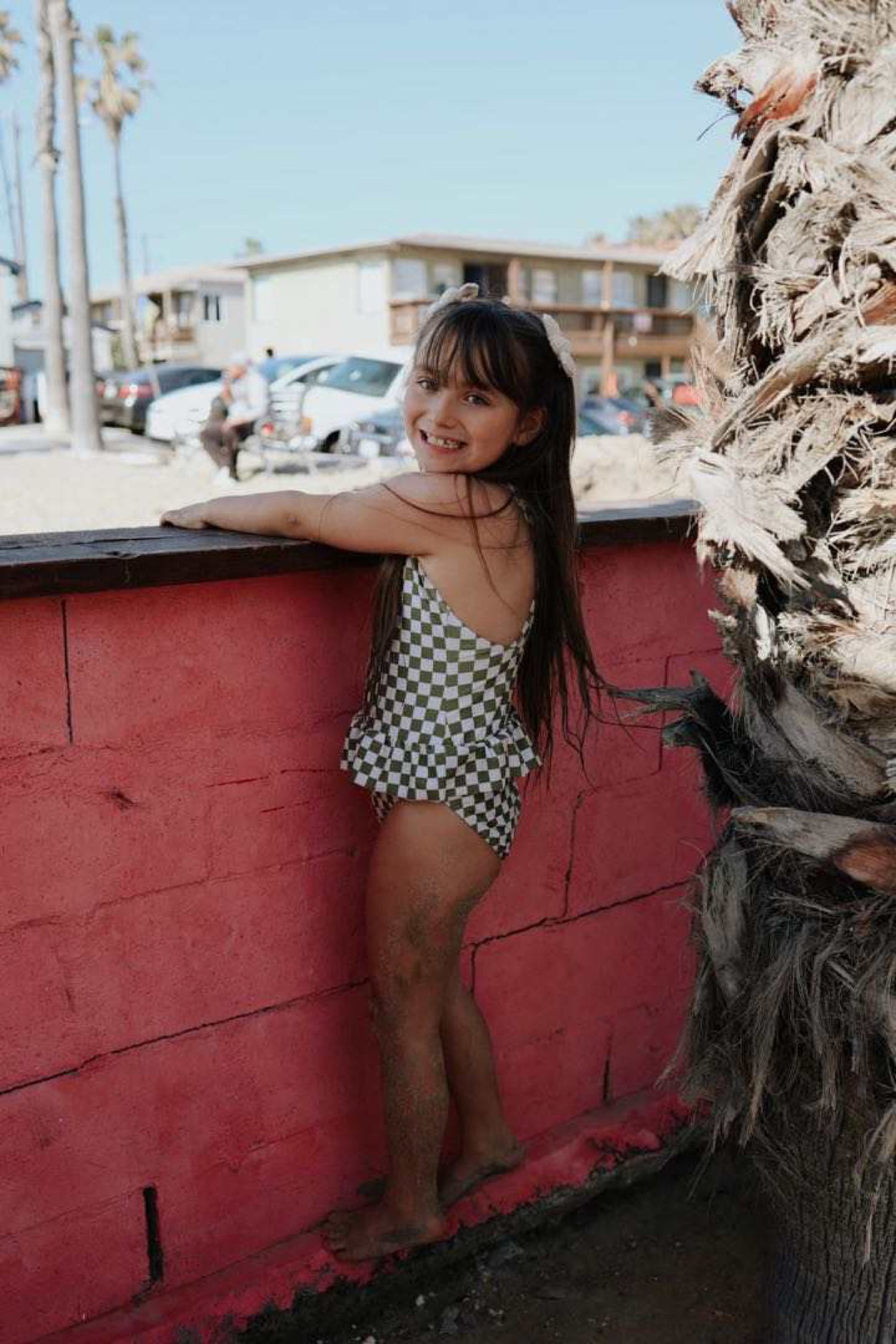 A young girl with long hair stands on a ledge against a red wall, smiling at the camera. She is barefoot and wearing the Wave Chaser Olive Checker one piece girls swim suit by forever french baby. In the background, there are palm trees, buildings, and parked cars under a clear blue sky.