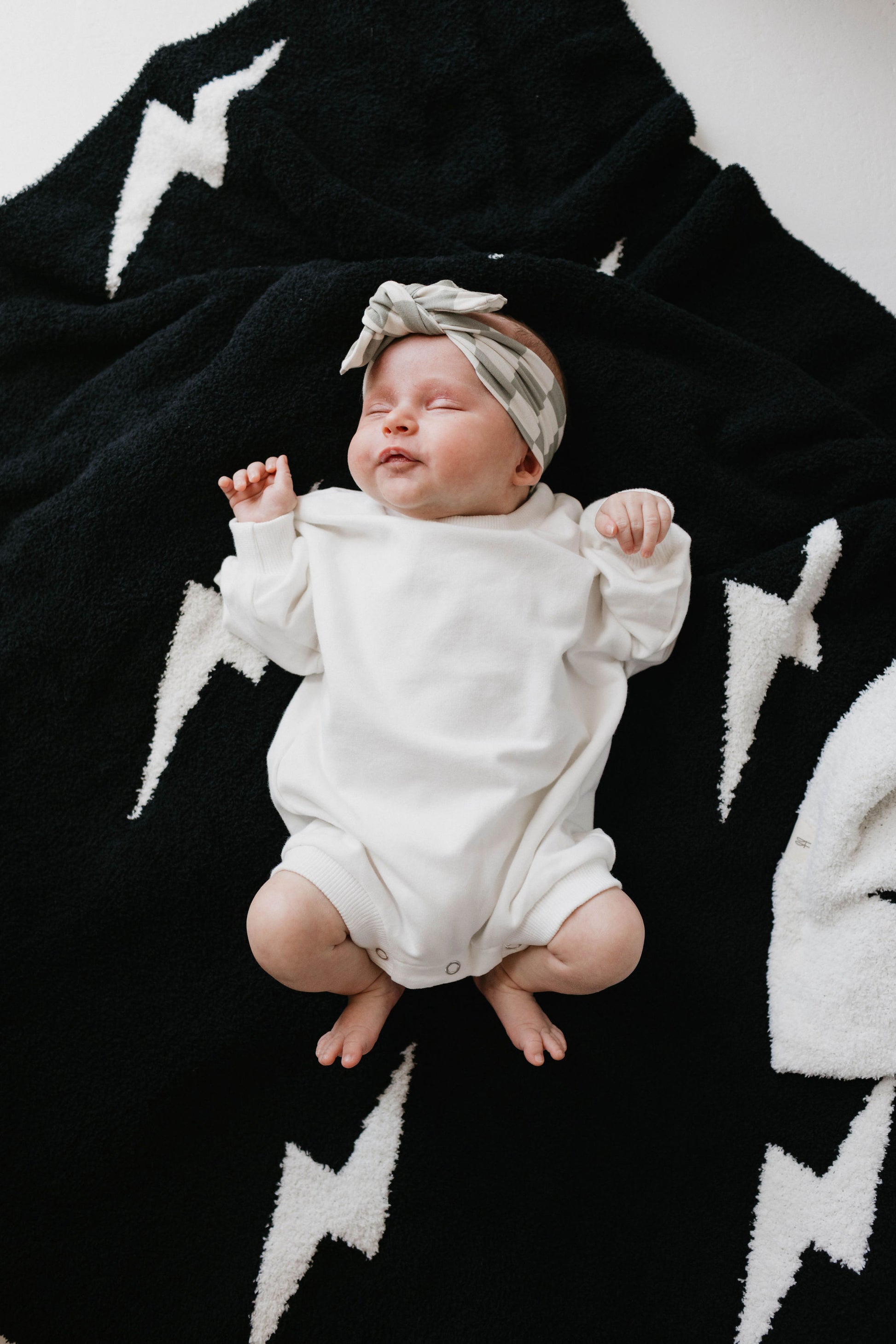 A baby wearing a Dove-colored Sweatshirt Romper and a plaid headband from Forever French Baby lies on a black blanket adorned with white lightning bolt patterns. The baby's eyes are closed, and their arms and legs are slightly raised. The scene exudes coziness and tranquility, capturing a perfect Forever French Baby moment.