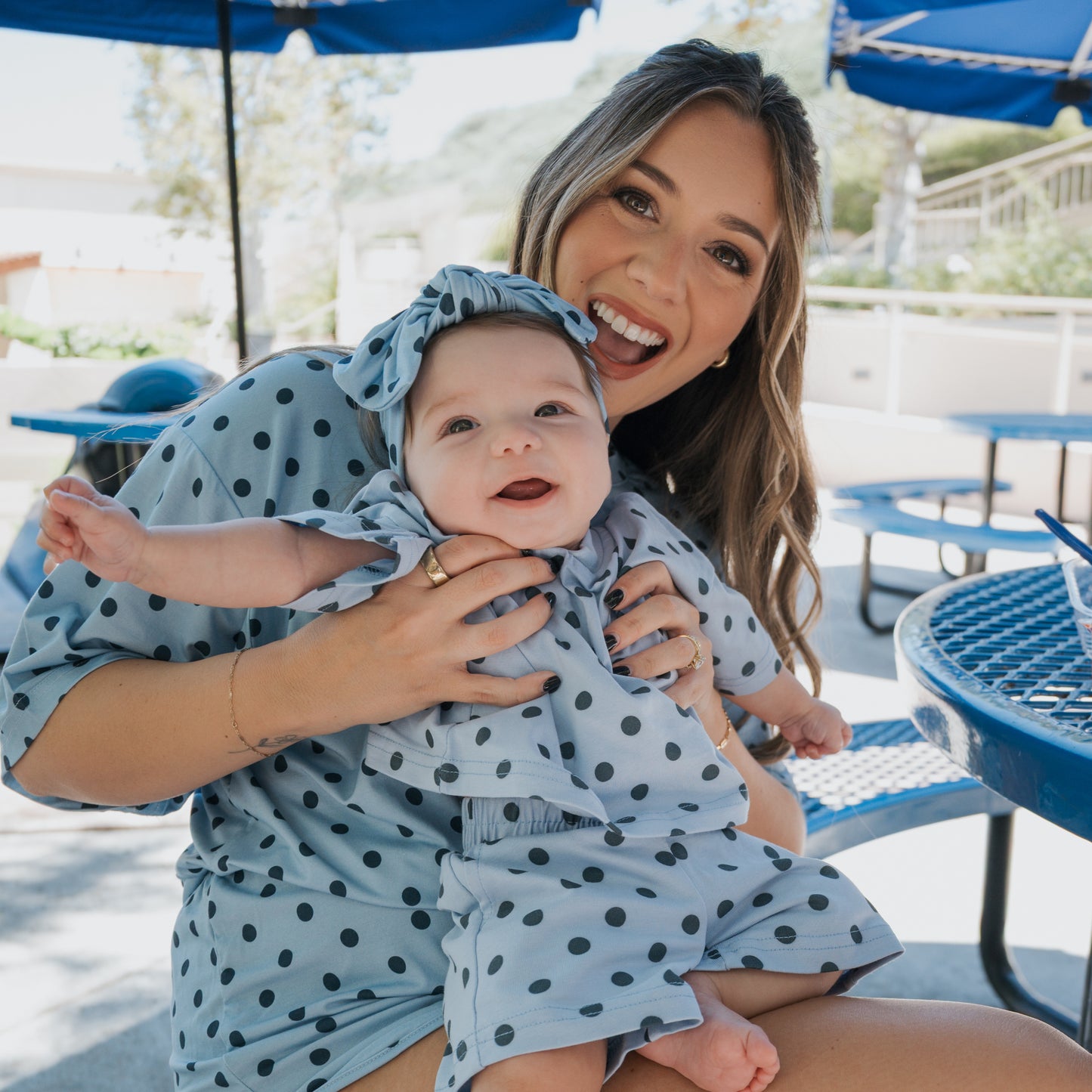 A smiling woman wearing the forever french baby Women's Bamboo Short Set | Sully in blue polka dots holds a happy baby dressed in matching hypo-allergenic bamboo clothing, as they sit together at a blue outdoor table on a sunny day.