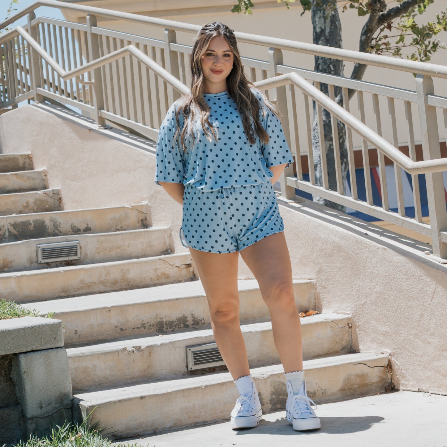 A woman stands on outdoor stairs, smiling and wearing the Women's Bamboo Short Set | Sully by forever french baby—a light blue matching polka dot pajama set featuring shorts and a loose top. She pairs the hypo-allergenic bamboo ensemble with white socks and white sneakers. The railings and building in the background highlight her relaxed, stylish look.