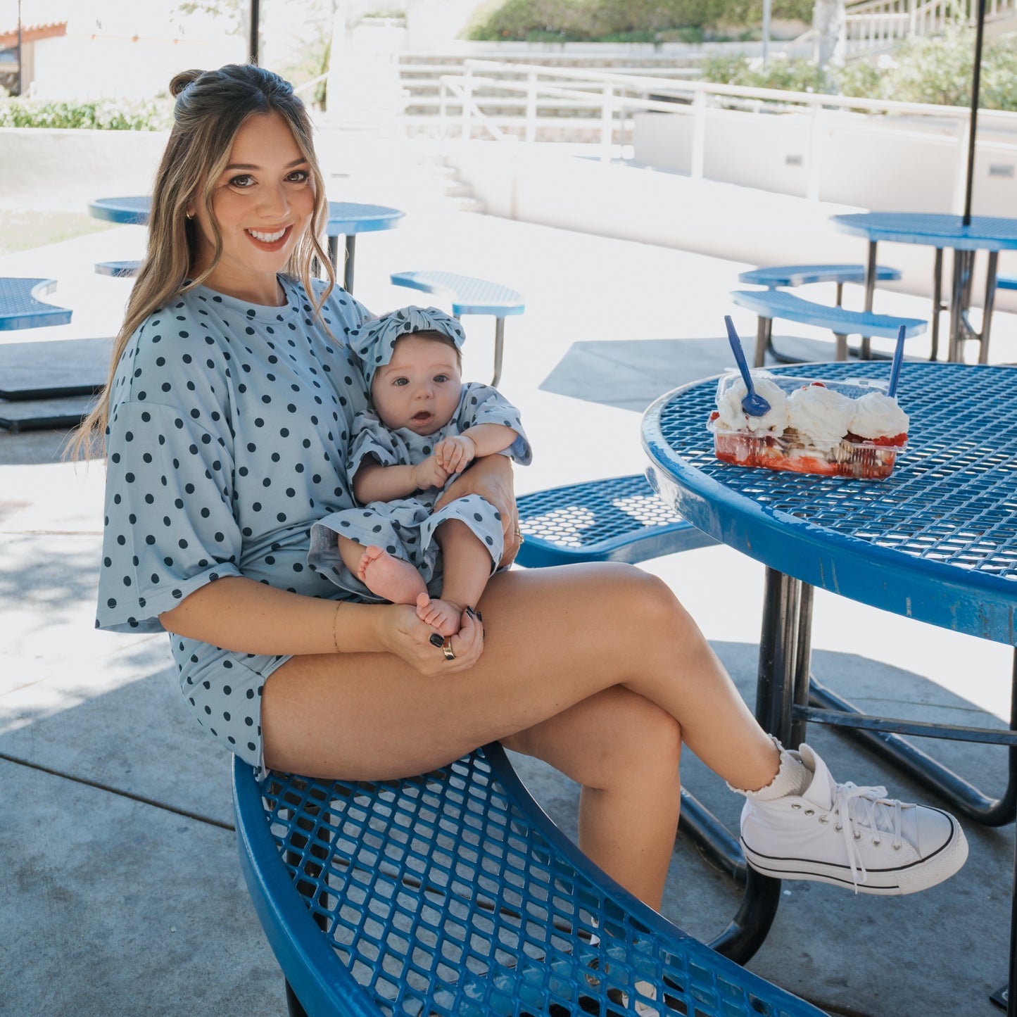 A woman wears the forever french baby Women's Bamboo Short Set | Sully in a blue polka-dot print while holding a matching baby, both seated outdoors at a blue picnic table with three frosted cupcakes and two spoons.