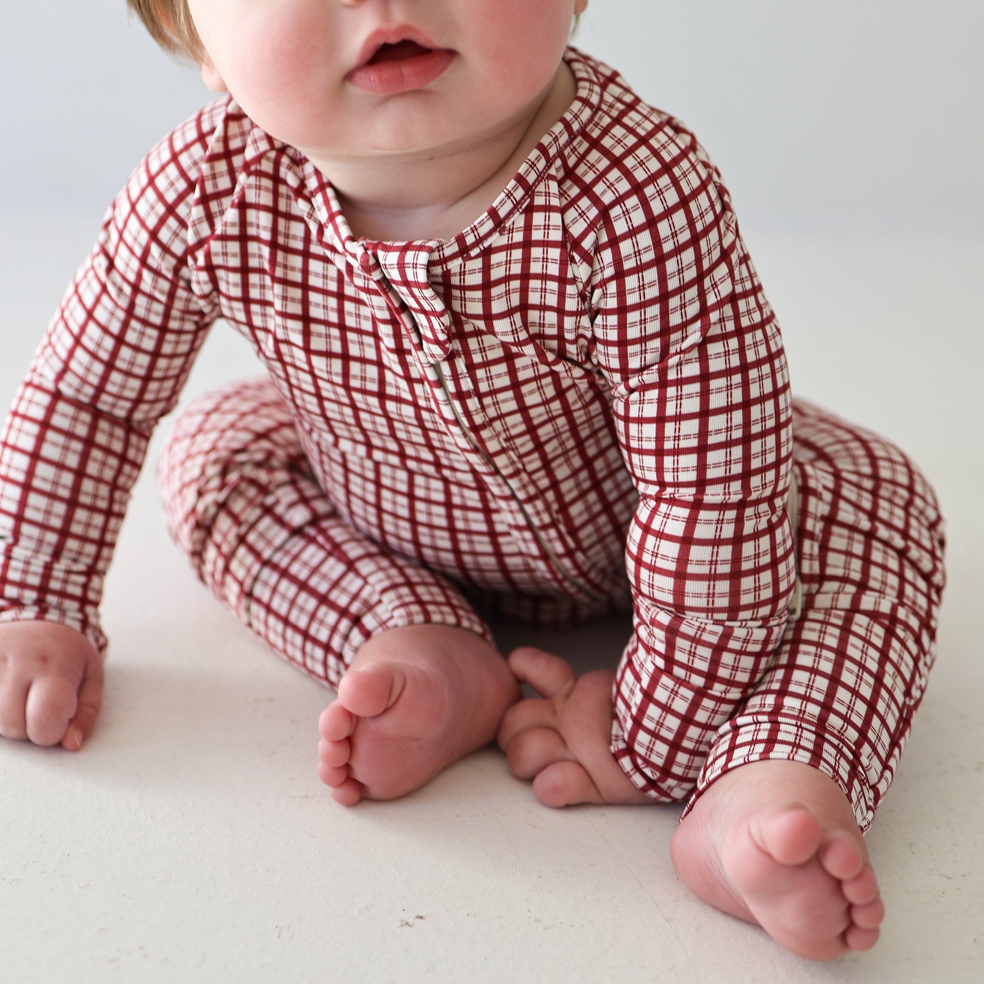 A baby sits on the floor wearing forever french baby's Bamboo Zip Pajamas in Window Pane. The baby's soft cheeks and lips are partly visible, with hands and bare feet gently resting on the floor.