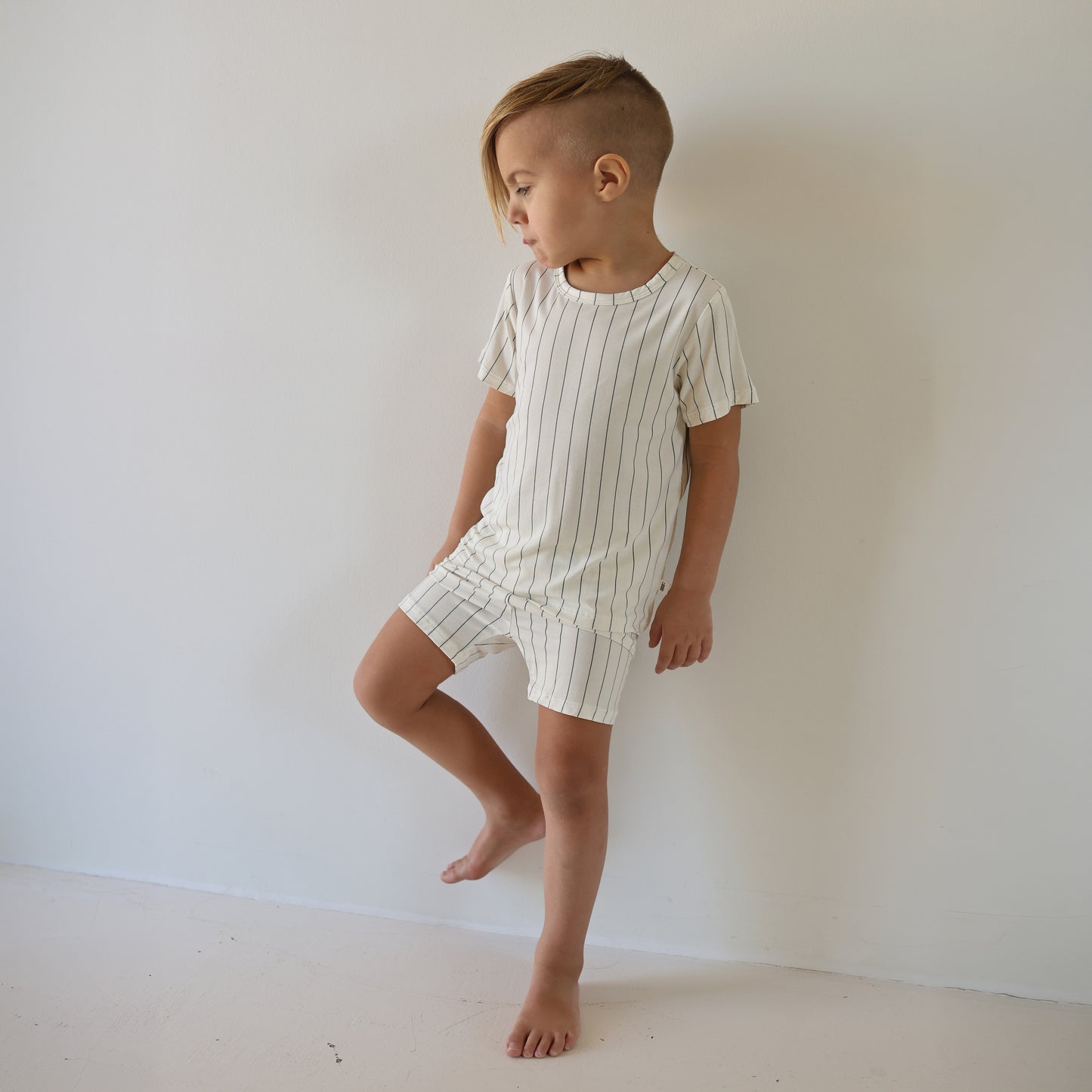 A young child with short, side-shaved hair stands barefoot against a white wall, wearing the forever french baby Bamboo Short Set | Jack—a breathable cream-colored kids’ outfit with thin black vertical stripes.
