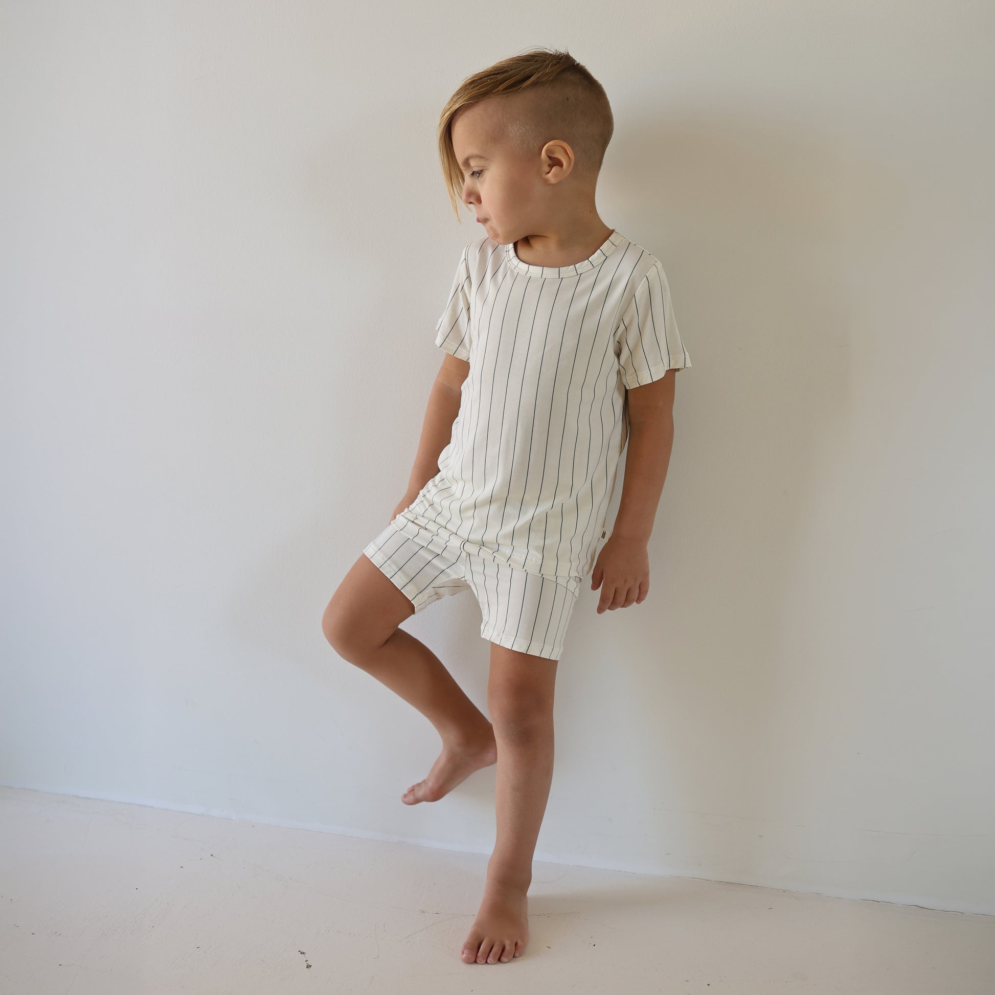 A young child with short, side-shaved hair stands barefoot against a white wall, wearing the forever french baby Bamboo Short Set | Jack—a breathable cream-colored kids’ outfit with thin black vertical stripes.