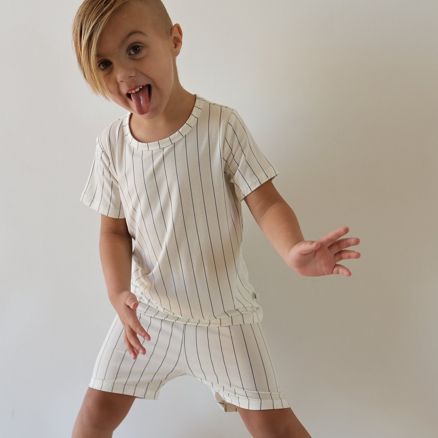 A young child with straight blonde hair and side-swept bangs poses playfully, wearing the Bamboo Short Set | Jack by forever french baby—a cream-colored, short-sleeve top and striped shorts—against a plain background.