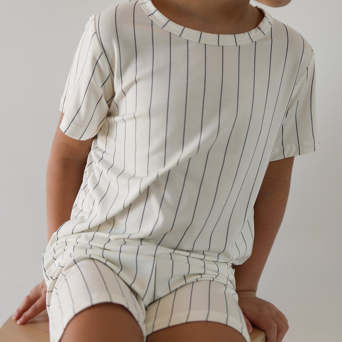 A child sits on a light surface, wearing the forever french baby Bamboo Short Set | Jack—a cream, short-sleeved kids outfit with thin navy blue stripes. The child’s face is partially hidden.