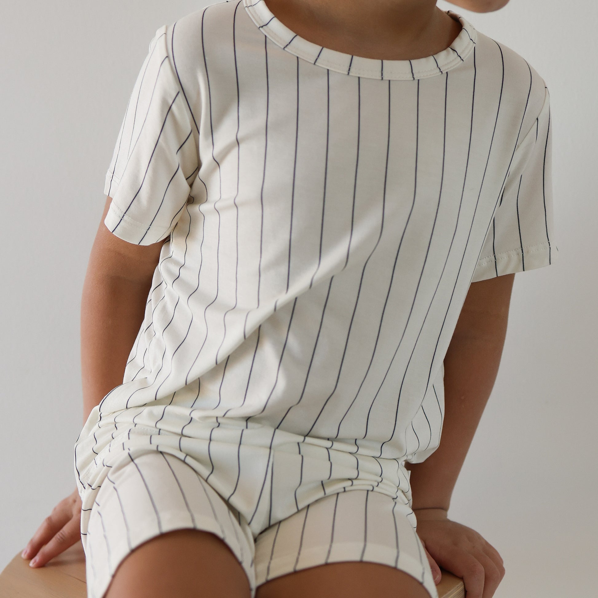 A child sits on a light surface, wearing the forever french baby Bamboo Short Set | Jack—a cream, short-sleeved kids outfit with thin navy blue stripes. The child’s face is partially hidden.