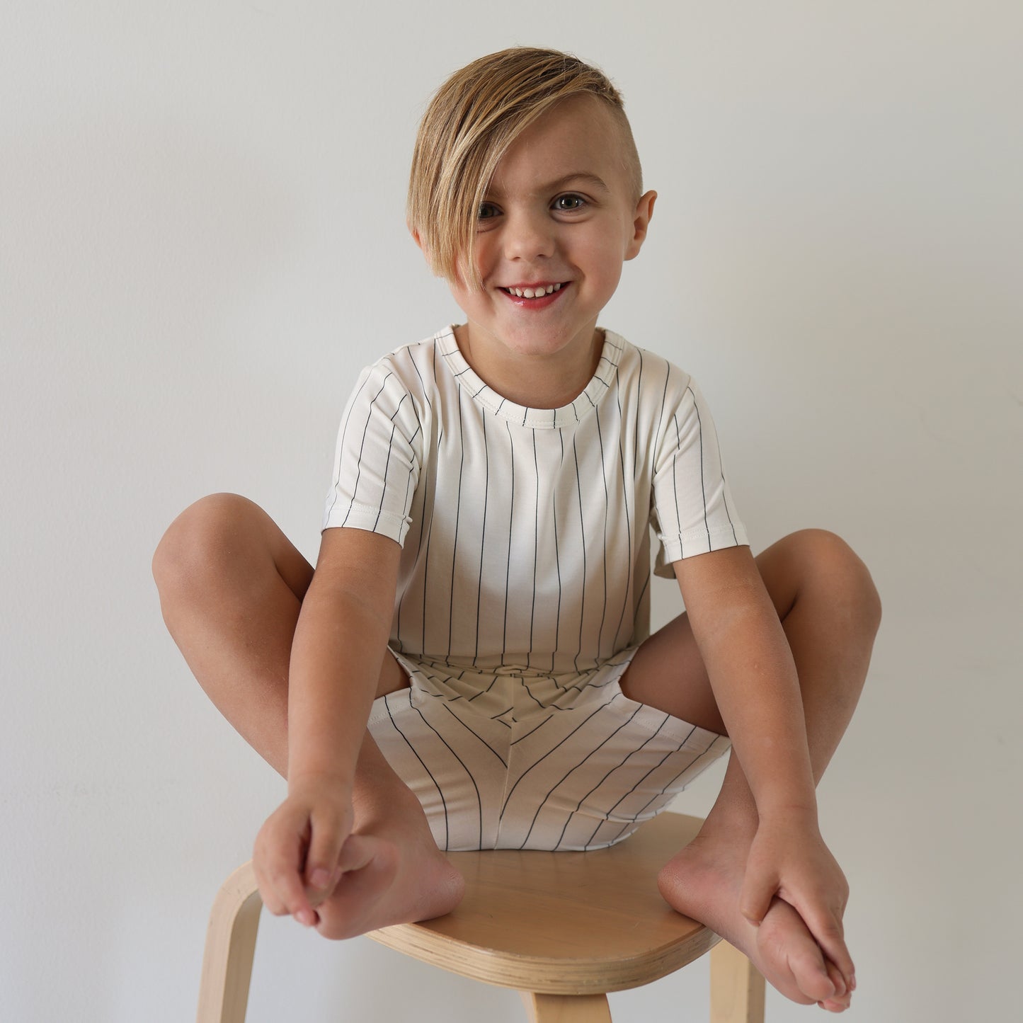 A smiling child with short, side-swept hair sits barefoot on a wooden stool, wearing the Bamboo Short Set | Jack by forever french baby—a white shirt with black vertical stripes—against a plain white background.