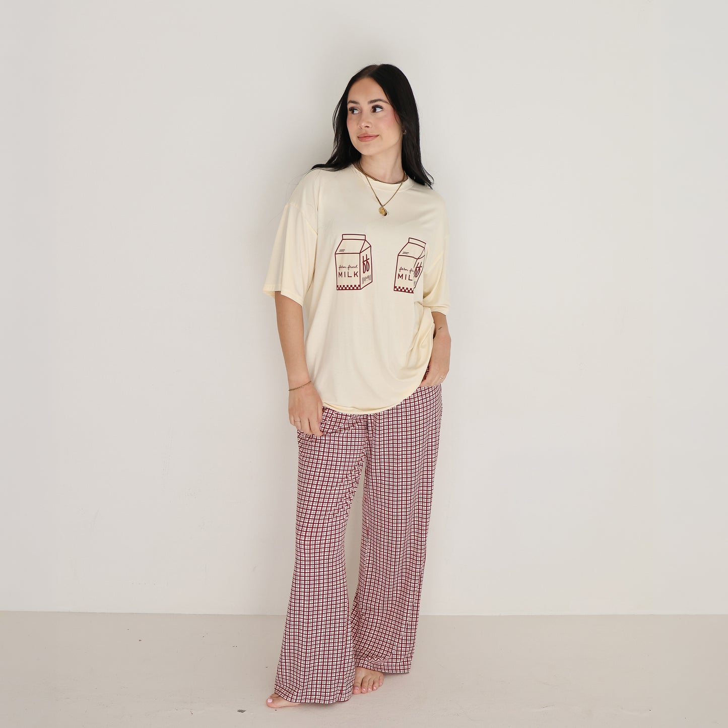 A woman with long dark hair stands barefoot against a white wall, wearing the forever french baby Short Sleeve Women's Bamboo Lounge Set in Milk & Window Pane and smiling softly as she looks to the side.