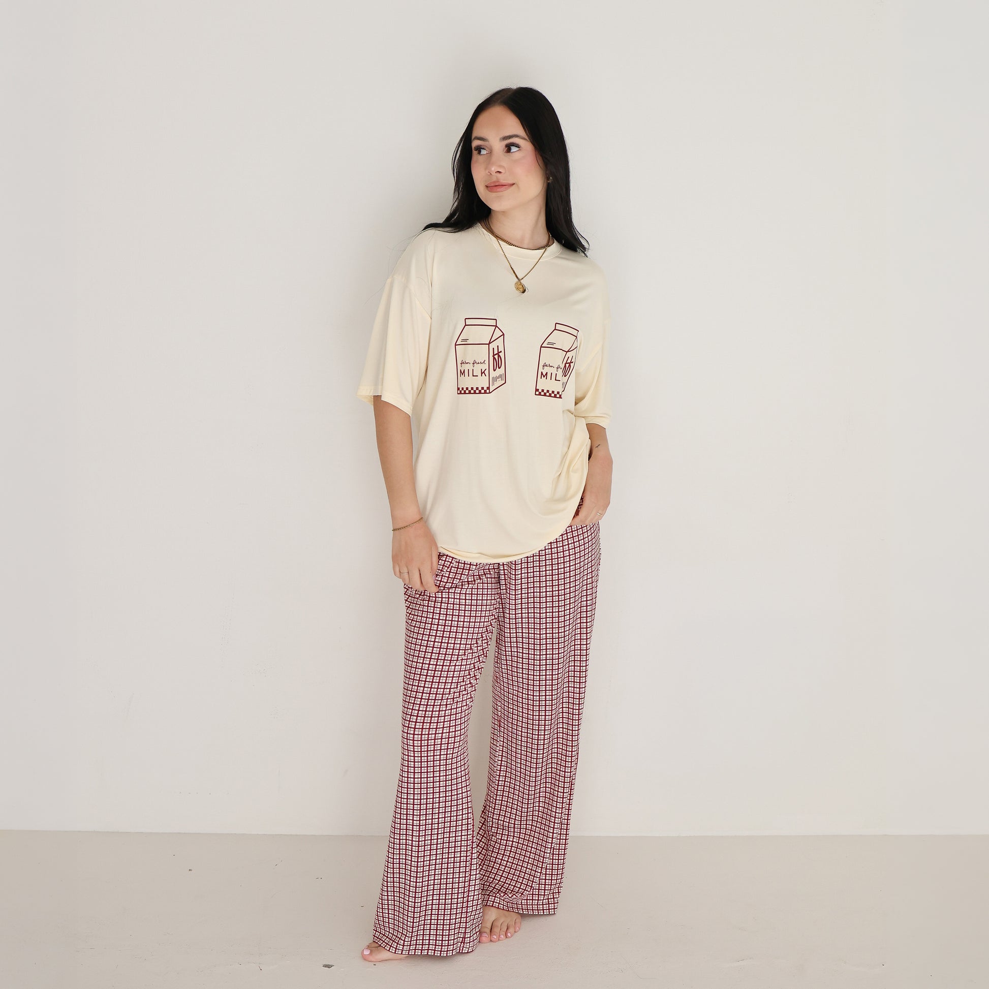 A woman with long dark hair stands barefoot against a white wall, wearing the forever french baby Short Sleeve Women's Bamboo Lounge Set in Milk & Window Pane and smiling softly as she looks to the side.
